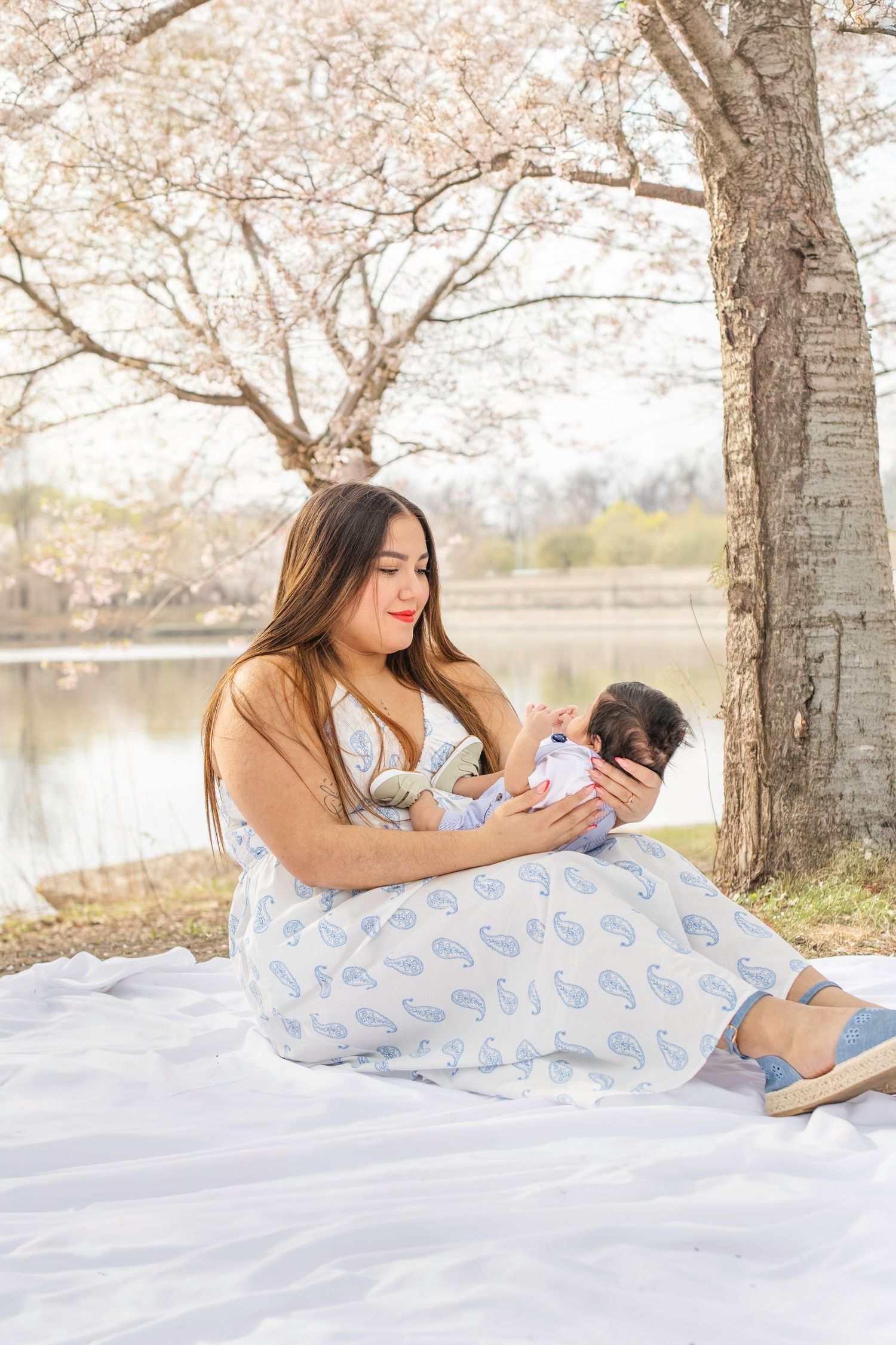 A tranquil scene of parent and baby relaxing on a white blanket under blooming cherry trees beside a lake.