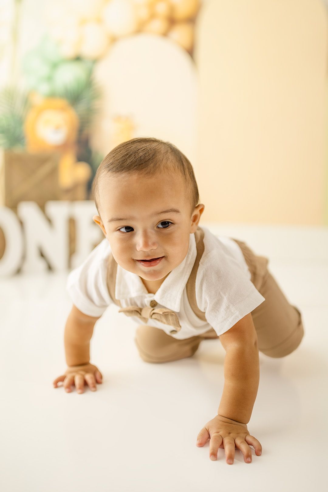 A smiling baby boy in a white shirt and tan suspenders crawls on a white floor at a first birthday party.