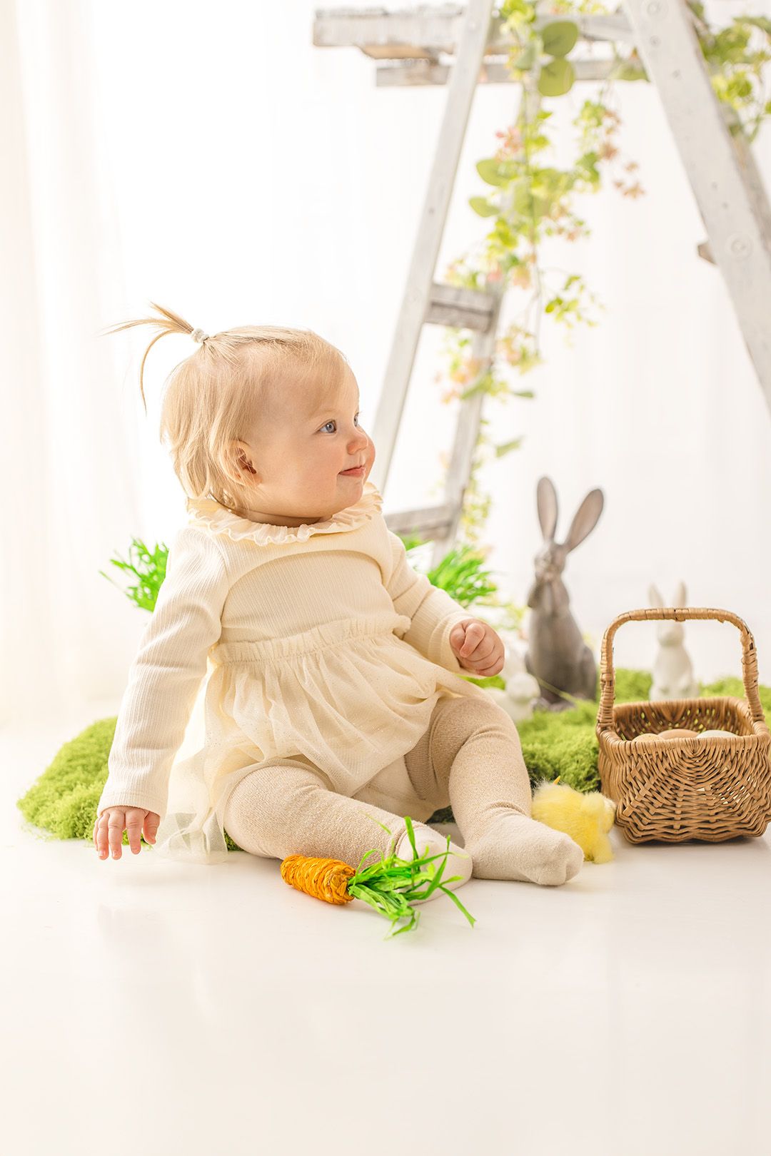 Baby in cream-colored outfit sits on white floor with Easter decorations including baskets and plush carrots.