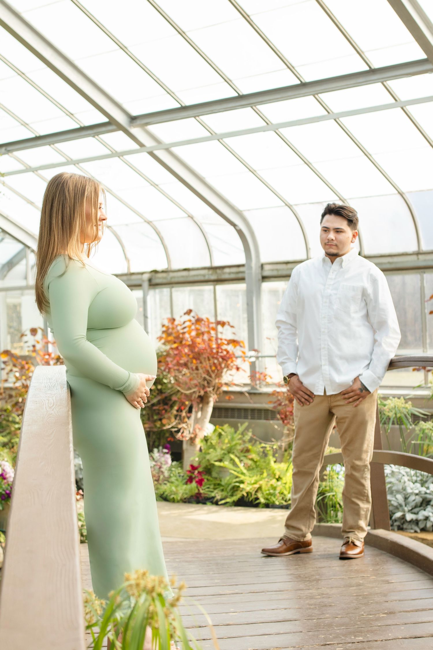 Maternity couple poses in a greenhouse with natural lighting and lush plant backdrop.