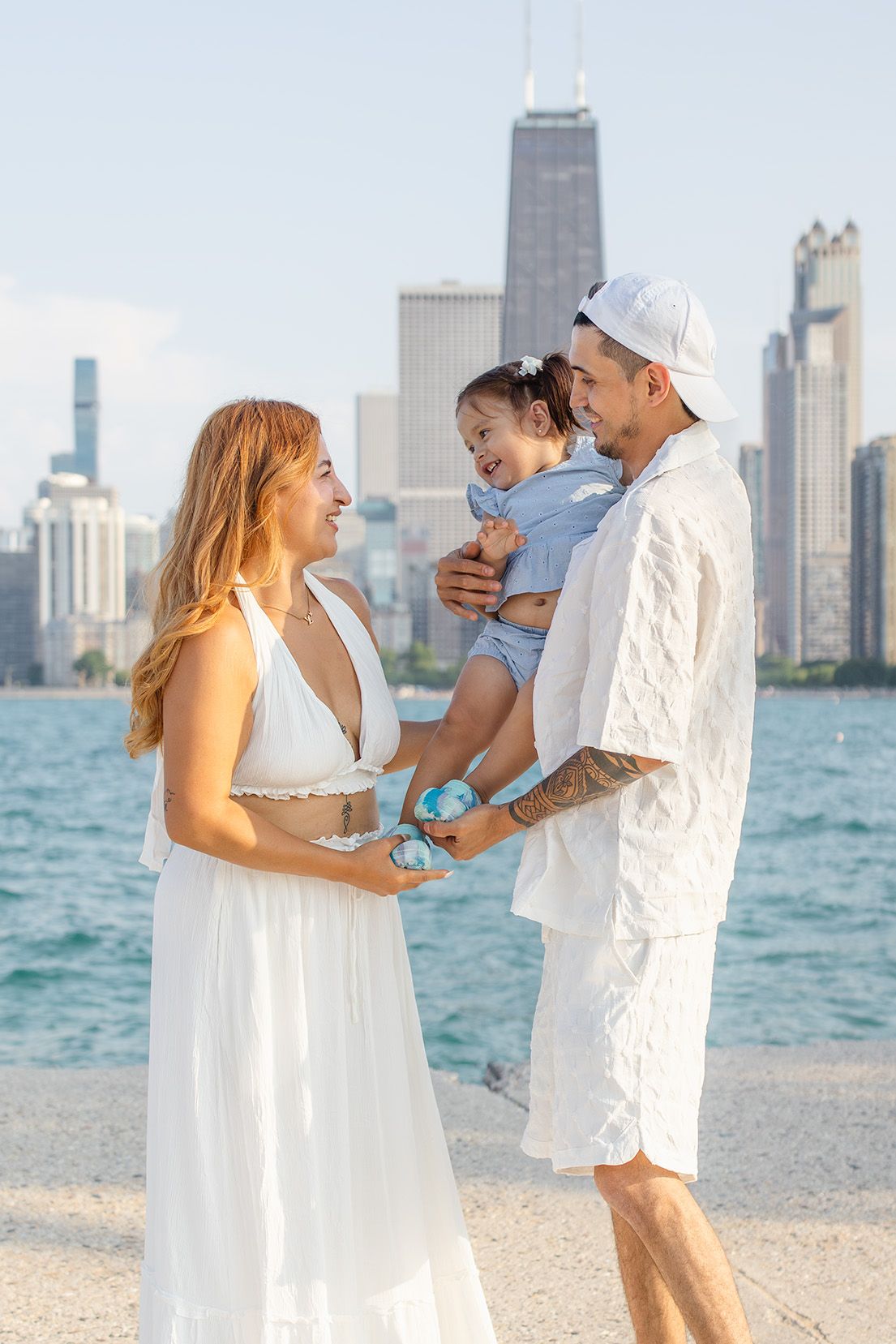 Happy family in white outfits poses with toddler by Chicago lakefront with city skyline in background.