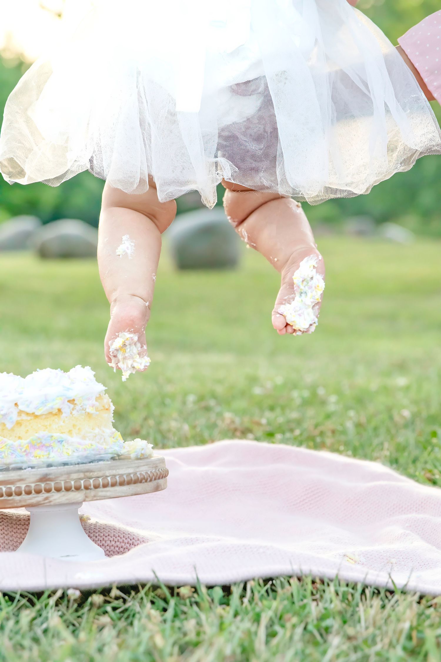 Baby's frosting-covered feet dangle above a smash cake during a first birthday outdoor celebration.