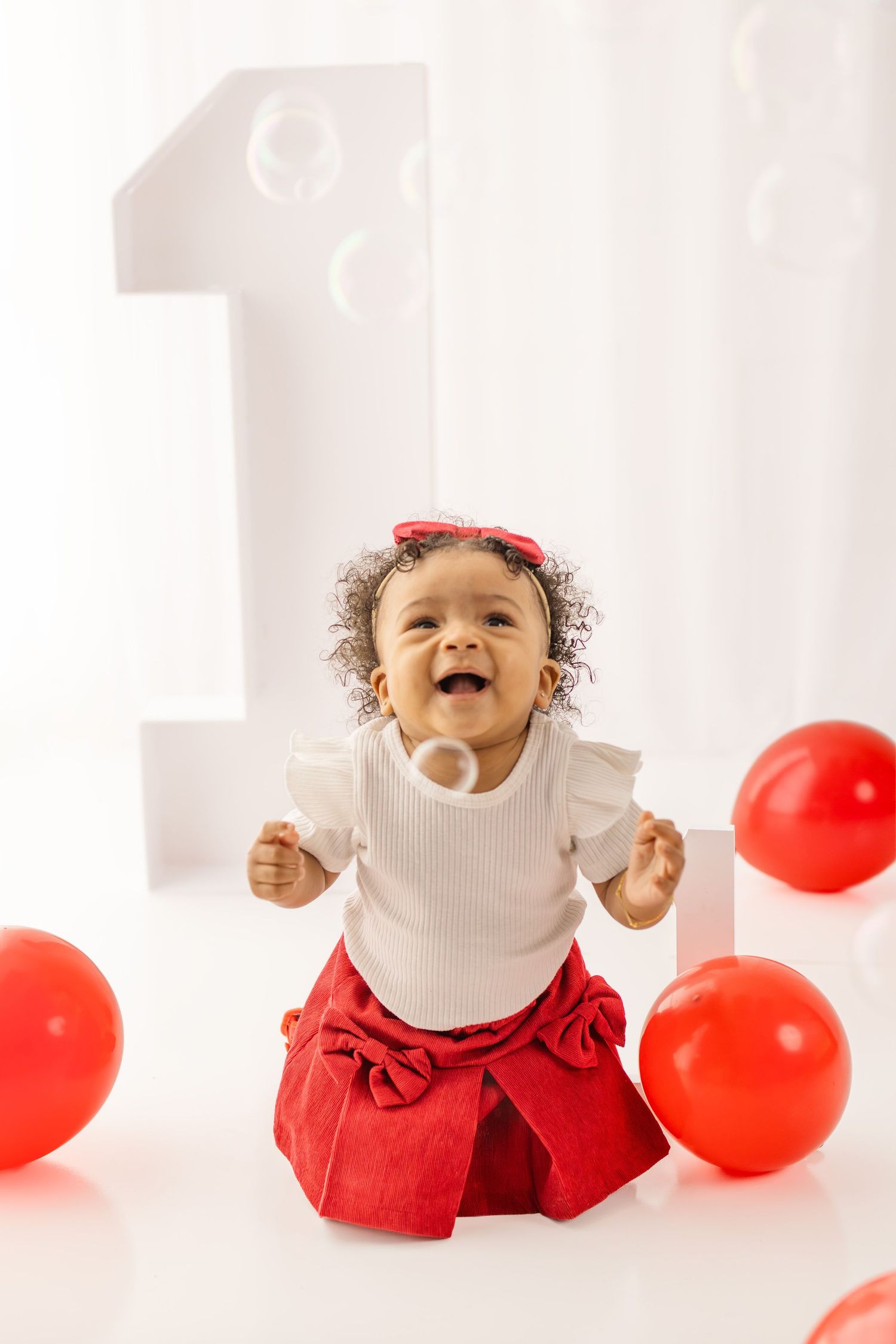 Joyful baby girl in red skirt and white top celebrates first birthday with red balloons and bubble.