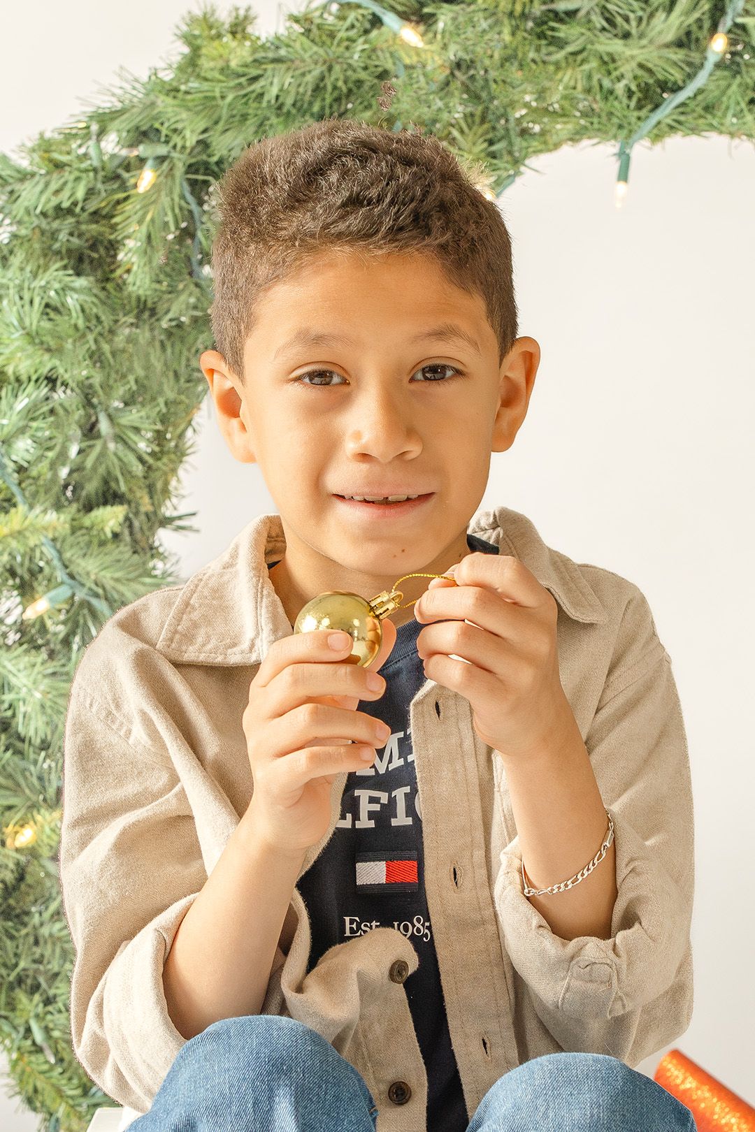 Young child in casual attire enjoying a cookie while sitting near evergreen branches during holiday season.