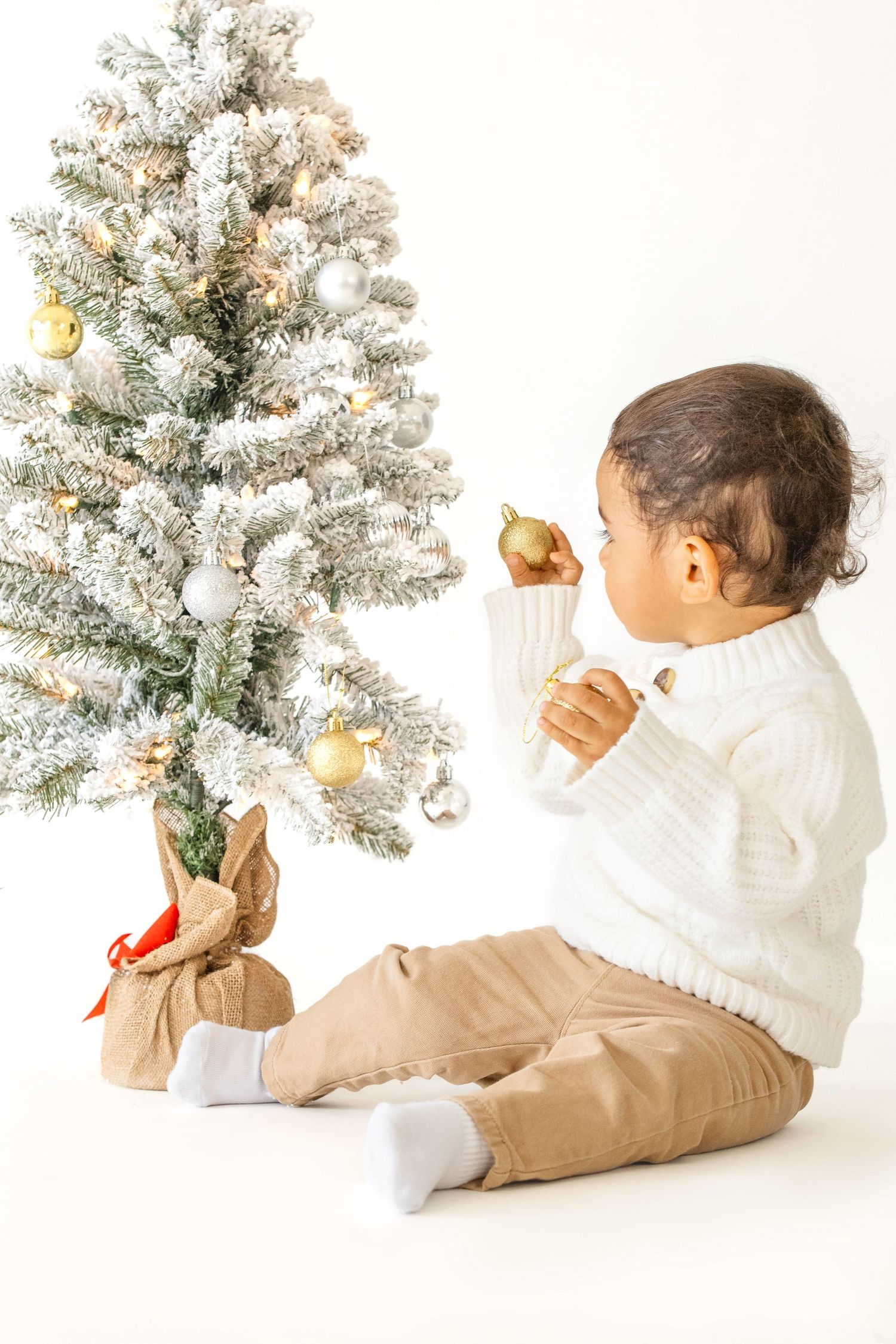 A baby in a white sweater and beige pants sits and plays with ornaments on a small frosted Christmas tree.