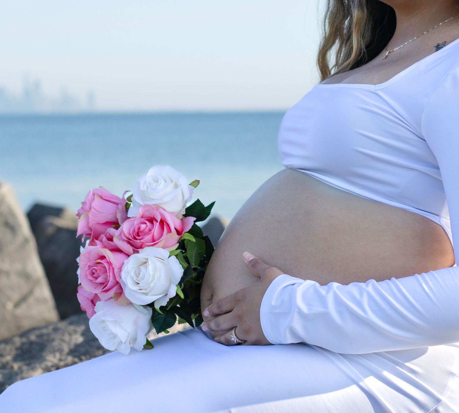 Close-up of pregnant belly with pink and white roses against ocean backdrop.