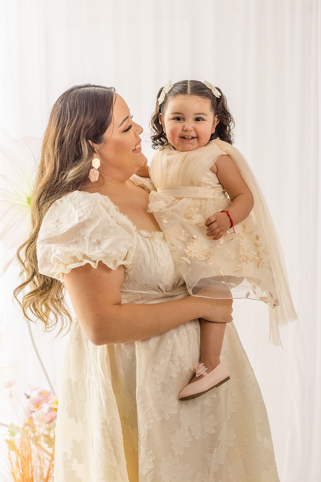 A mother in a cream lace dress holds her smiling toddler daughter, both wearing matching ivory outfits in a bright studio.