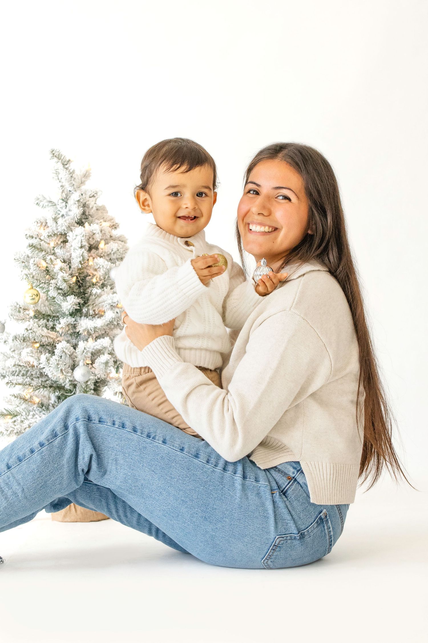 A parent and child wearing white sweaters lay on the floor together in front of a flocked Christmas tree.