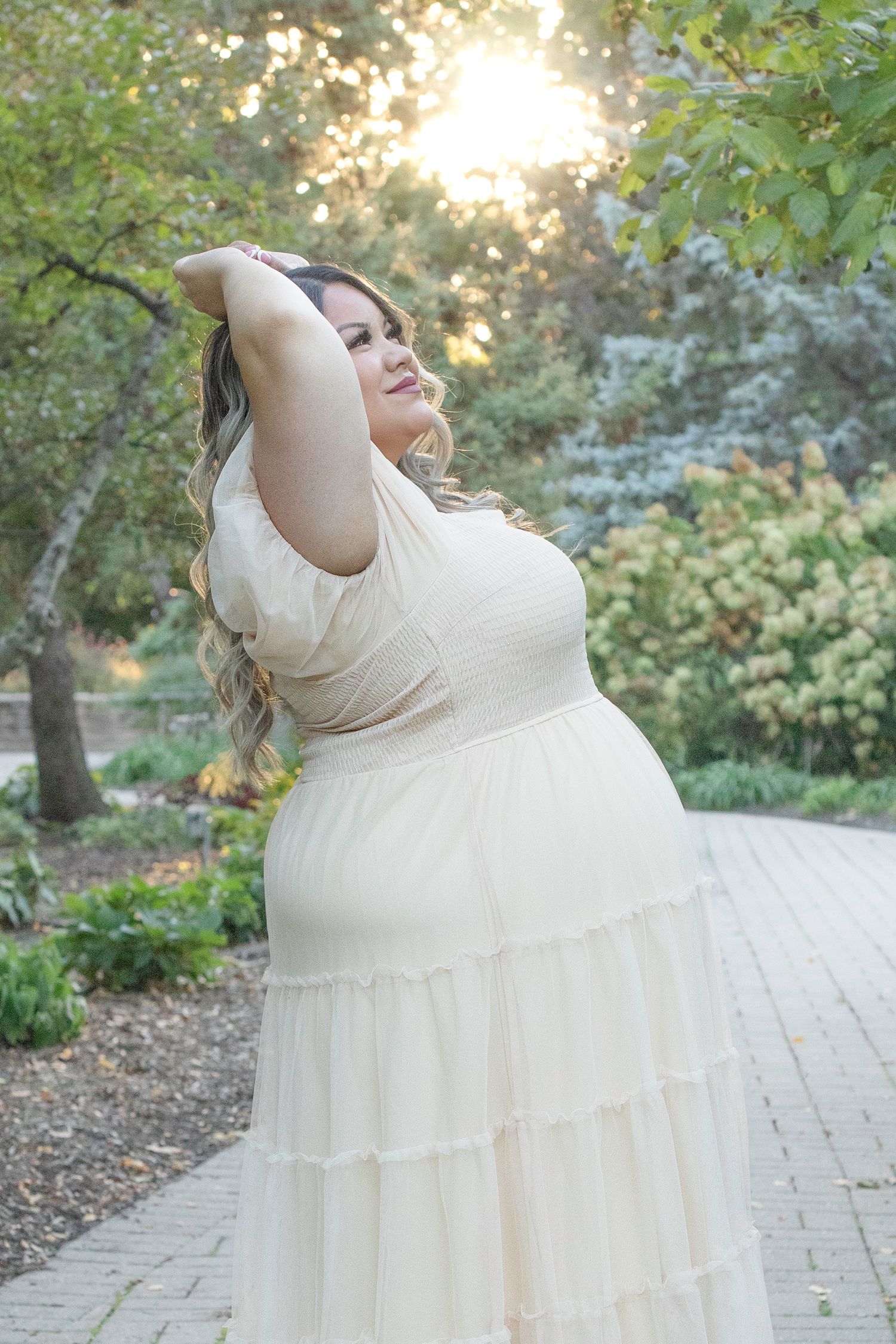 A person in a flowing white dress twirls joyfully on a stone path surrounded by lush greenery and sunlit trees.