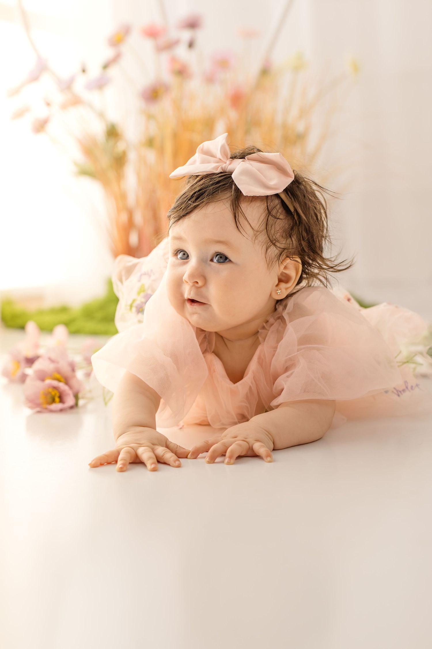 A baby girl in a pink tulle dress and bow headband lies on her tummy, looking up curiously, surrounded by soft florals.