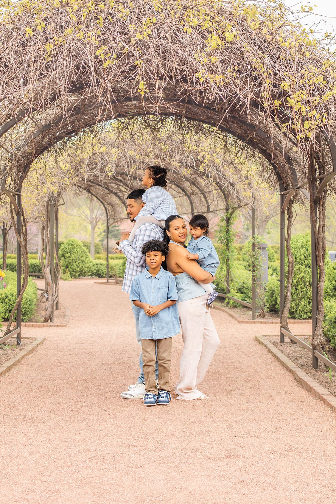 Family of four posing under a vine-covered garden archway, mother holding toddler with two boys in front.