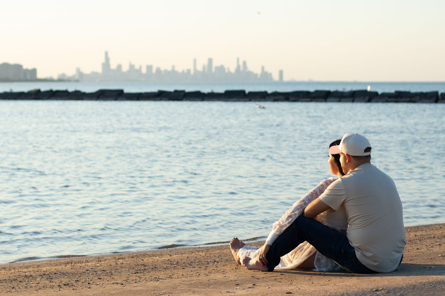 Person sits on rocky shoreline overlooking distant city skyline at sunset.