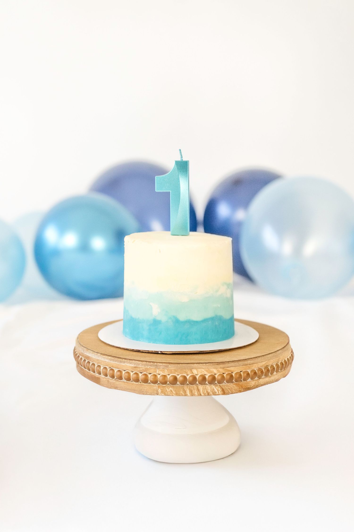 Blue ombre first birthday cake with number 1 candle on wooden stand, blue balloons in background.