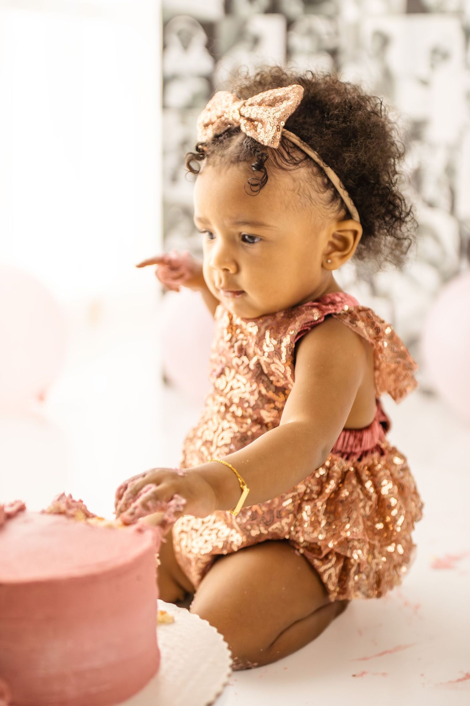 Adorable baby girl in gold sequin romper and bow headband smashing pink birthday cake during first birthday photoshoot.