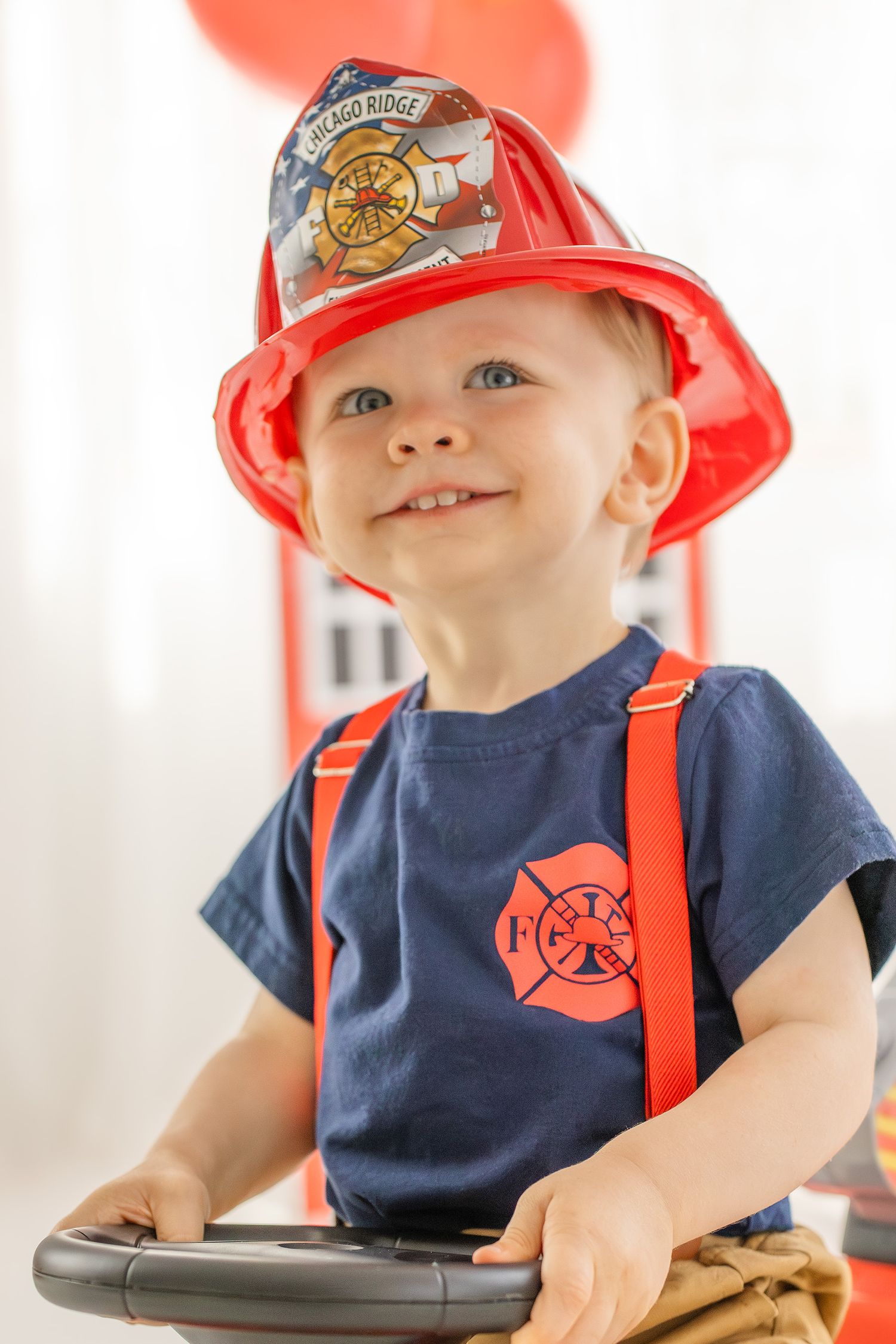 Cute toddler boy in firefighter costume with red helmet and suspenders smiles during birthday party.