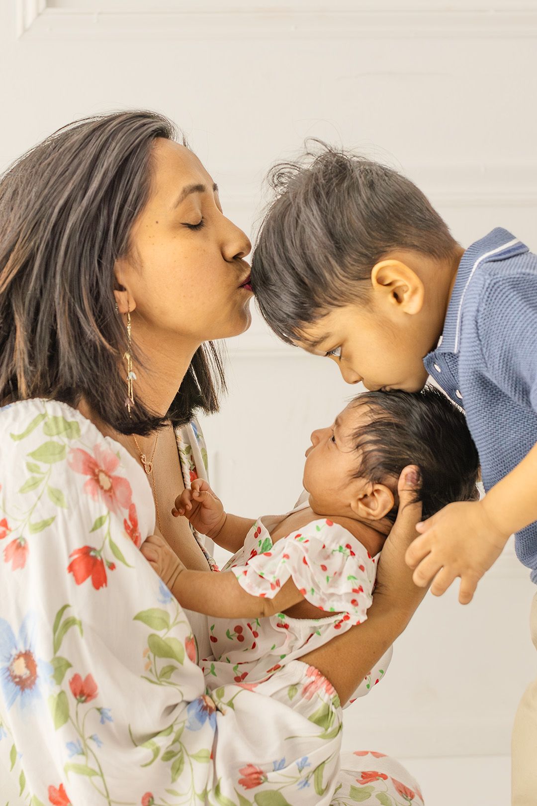 A mother kisses her young son while cradling her newborn baby, sharing a tender family moment.