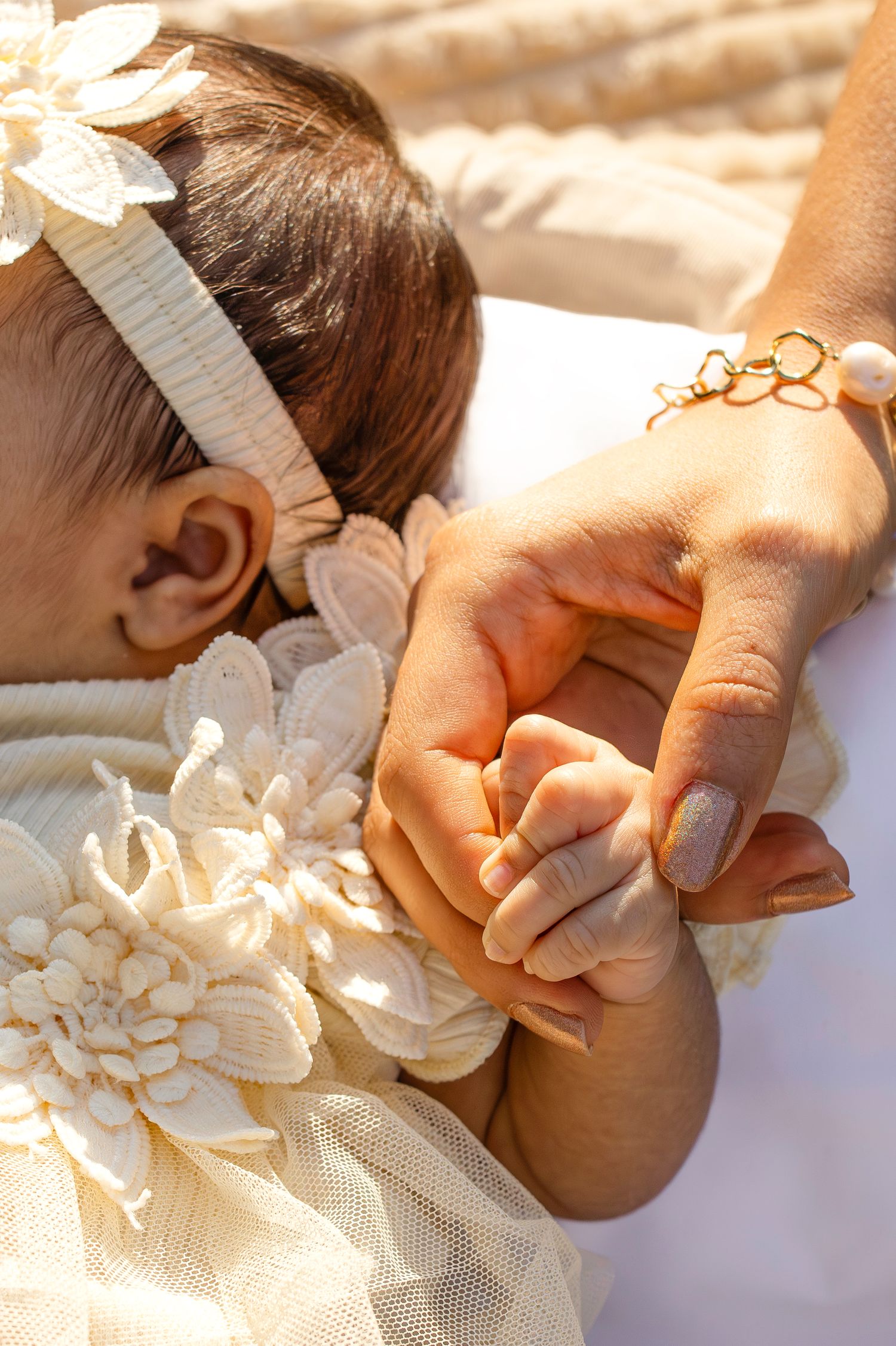A small baby hand holds onto an adult's finger while wearing a delicate white dress with floral details and hair bow.