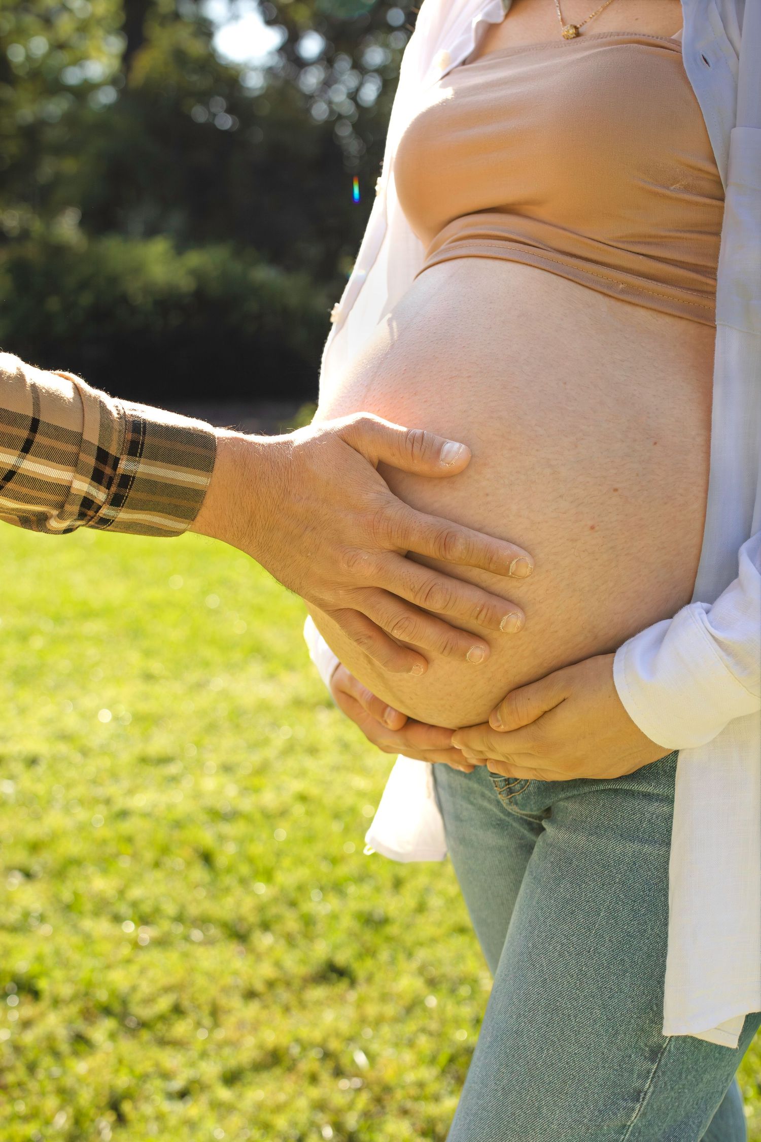 Close up series of hands gently cradling a pregnant belly in a sunny outdoor setting with green grass in background.