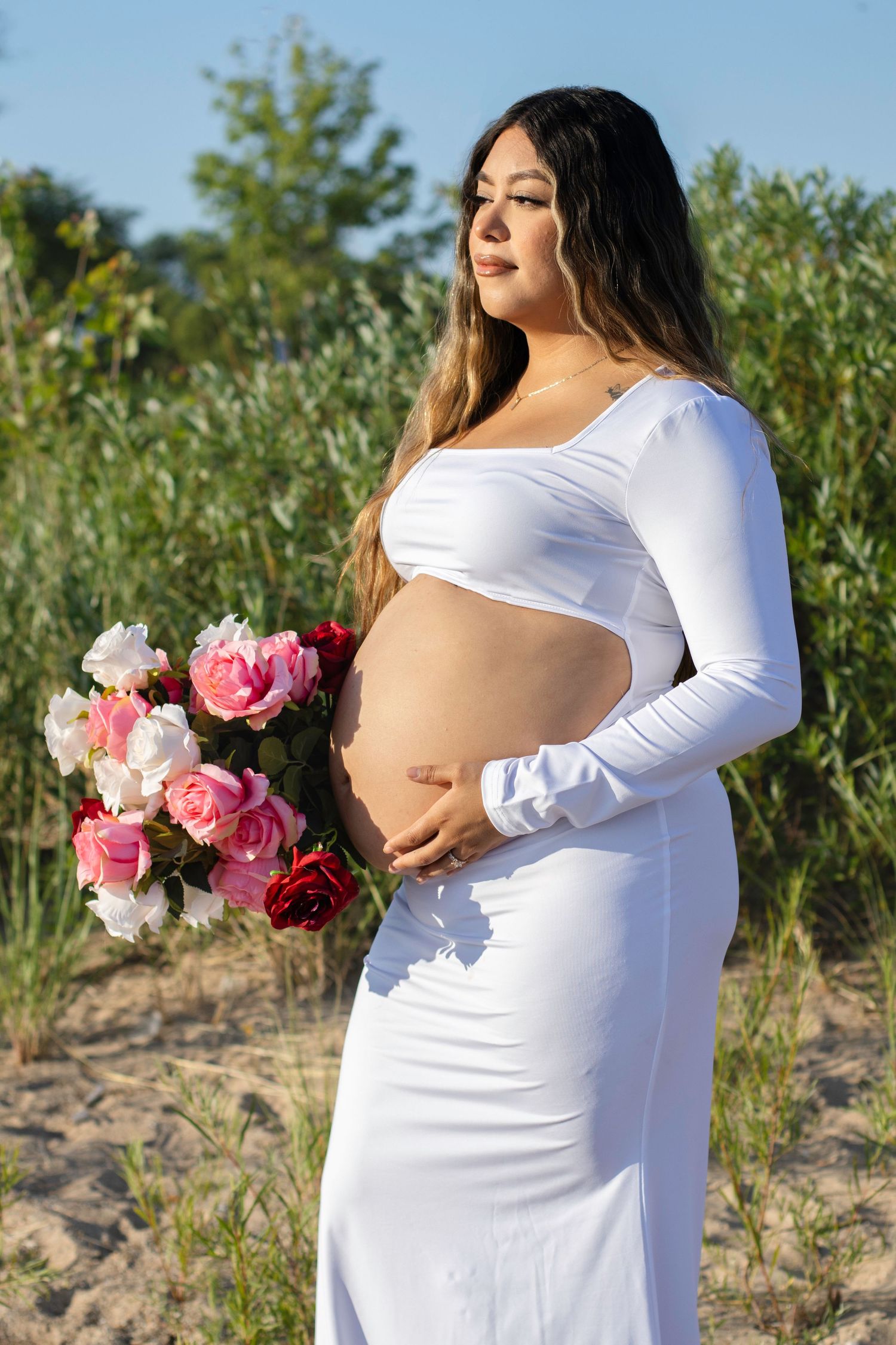 Pregnant woman in white dress holds pink roses during outdoor maternity photoshoot.