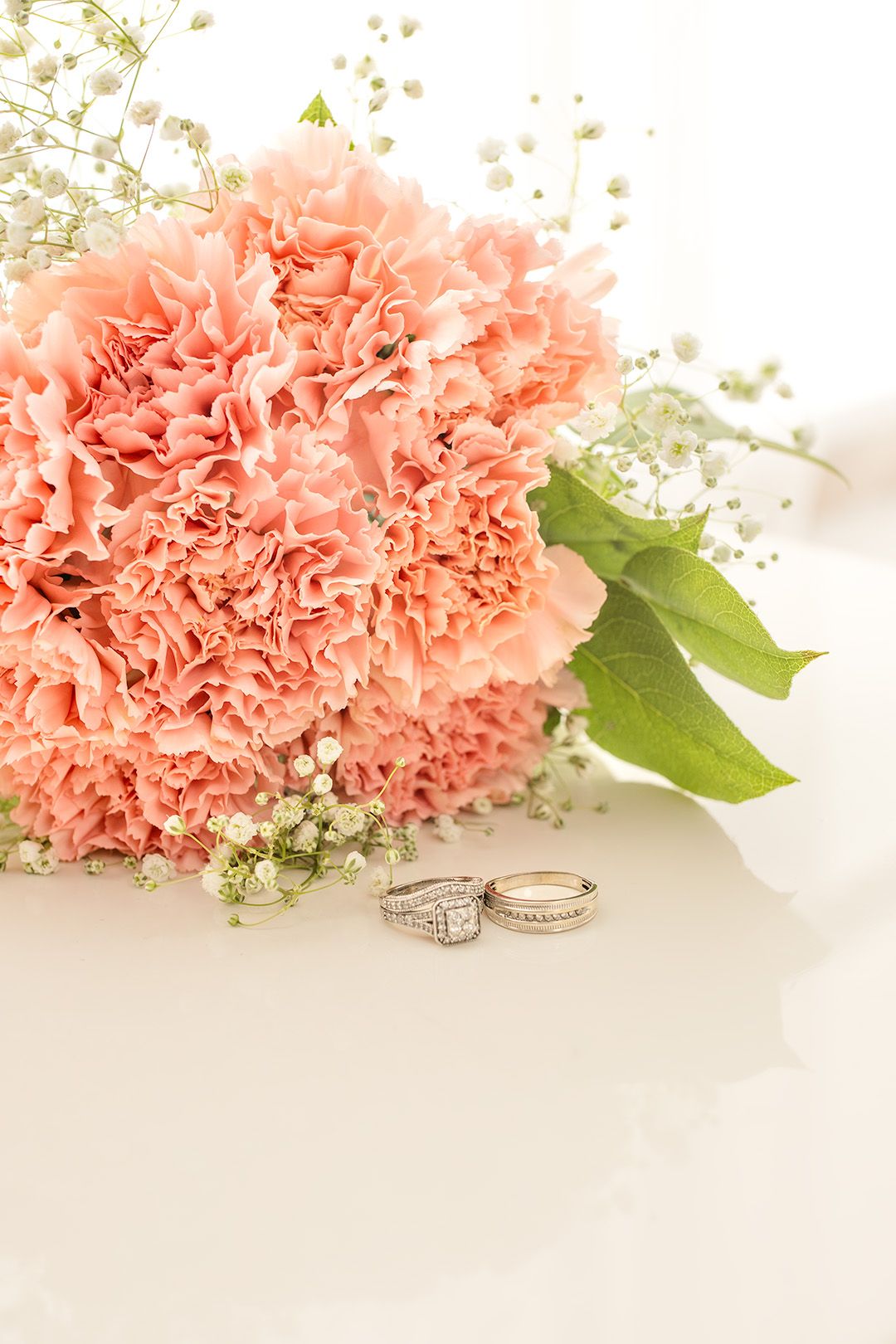 Pink carnation bouquet with baby's breath and two diamond wedding rings on a white surface.