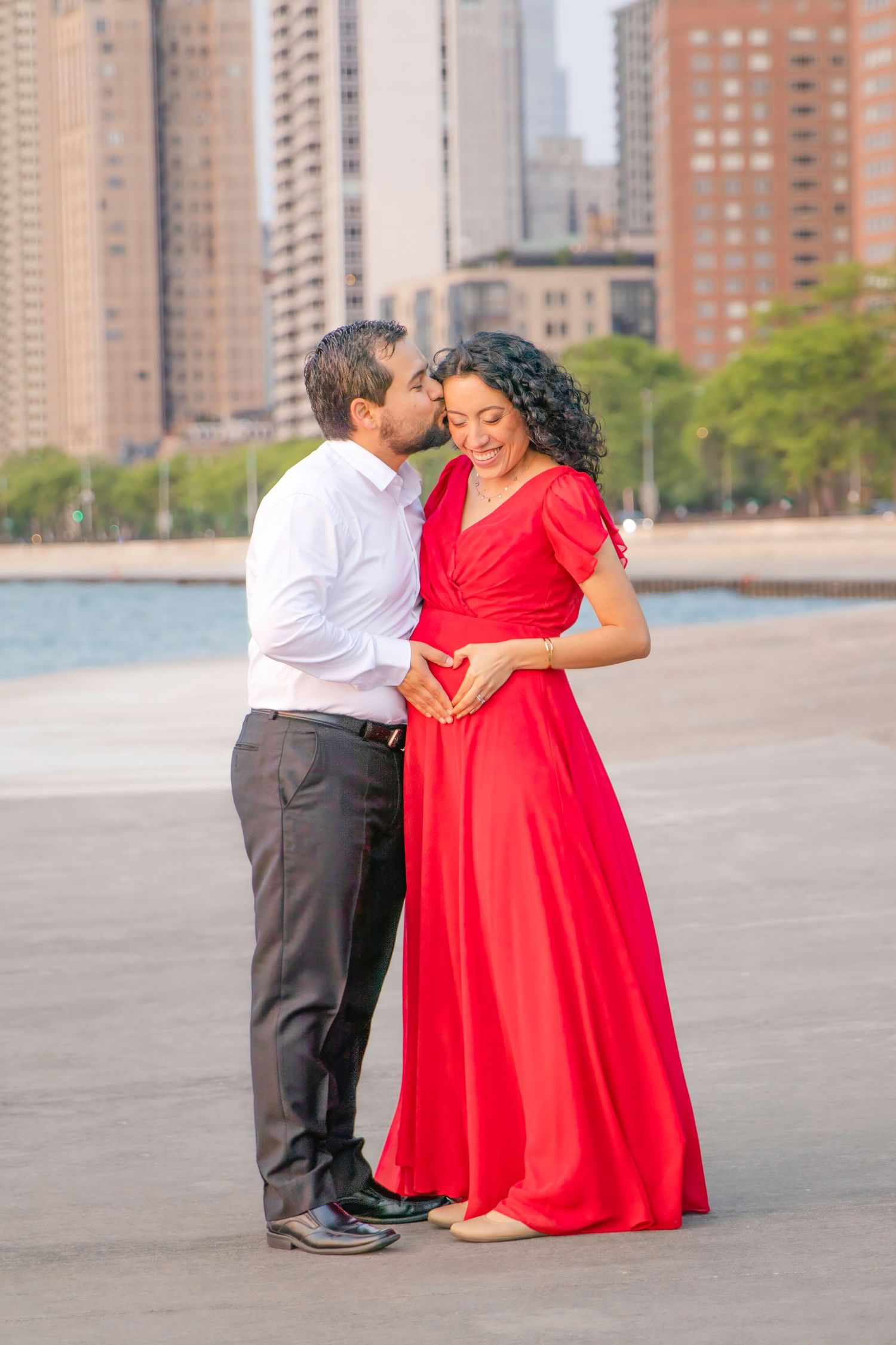 A couple shares a tender moment by the waterfront, with the woman wearing a flowing red dress and city buildings in the background.