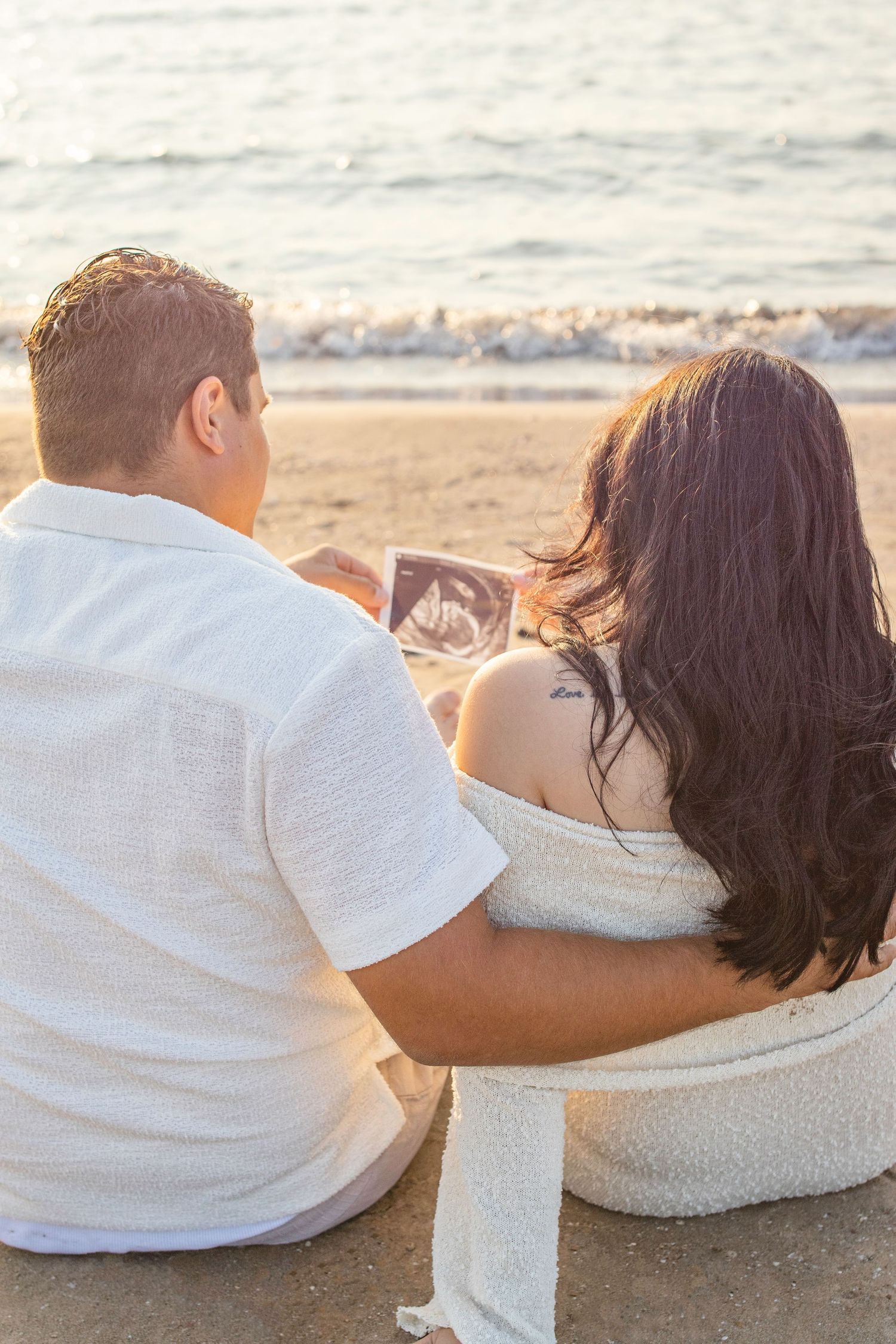 Couple sitting on beach looking at ultrasound photo together during golden hour by the ocean.