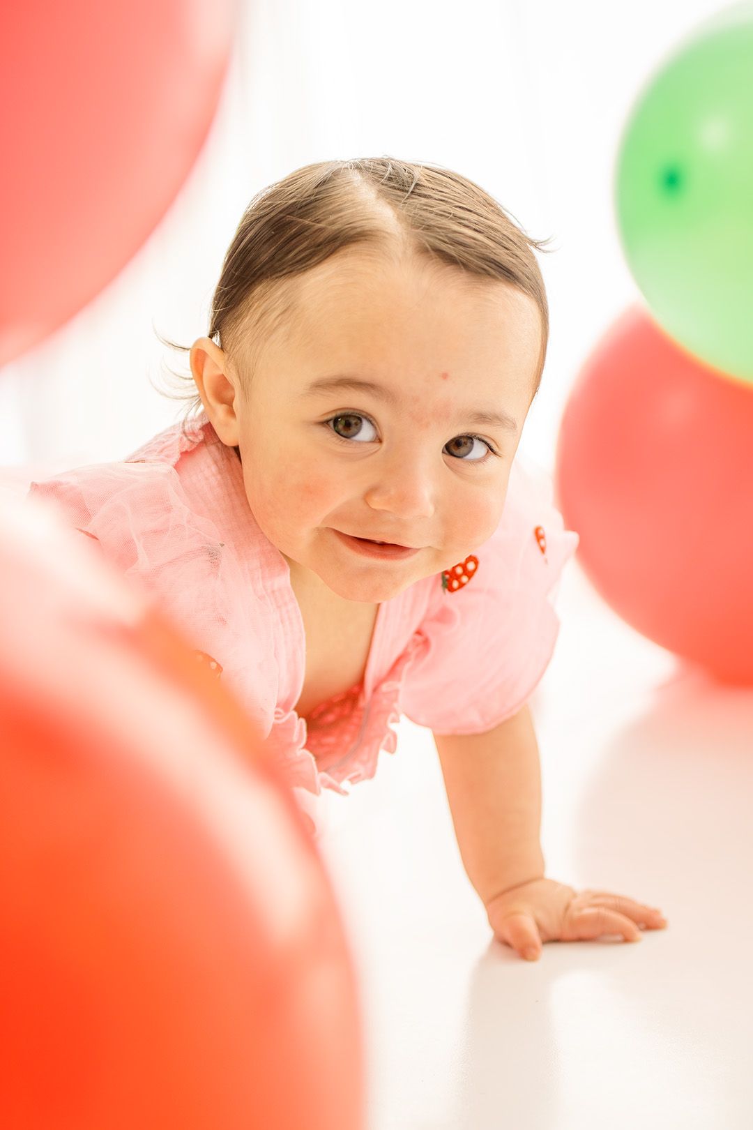 Baby girl in pink dress crawling among red and green balloons, smiling at camera during first birthday celebration.