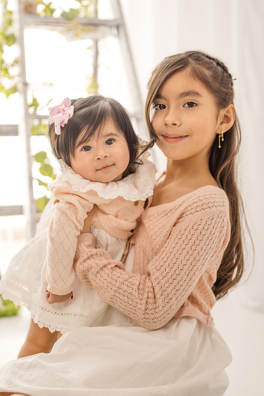 Young girl in pink sweater holds baby sister in white dress with floral hair clip in bright white room.