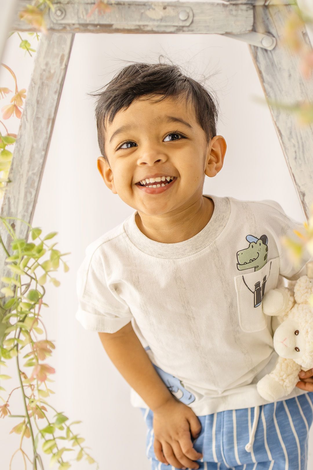 Smiling toddler boy in white graphic tee and blue striped shorts, posed near a rustic ladder with greenery.