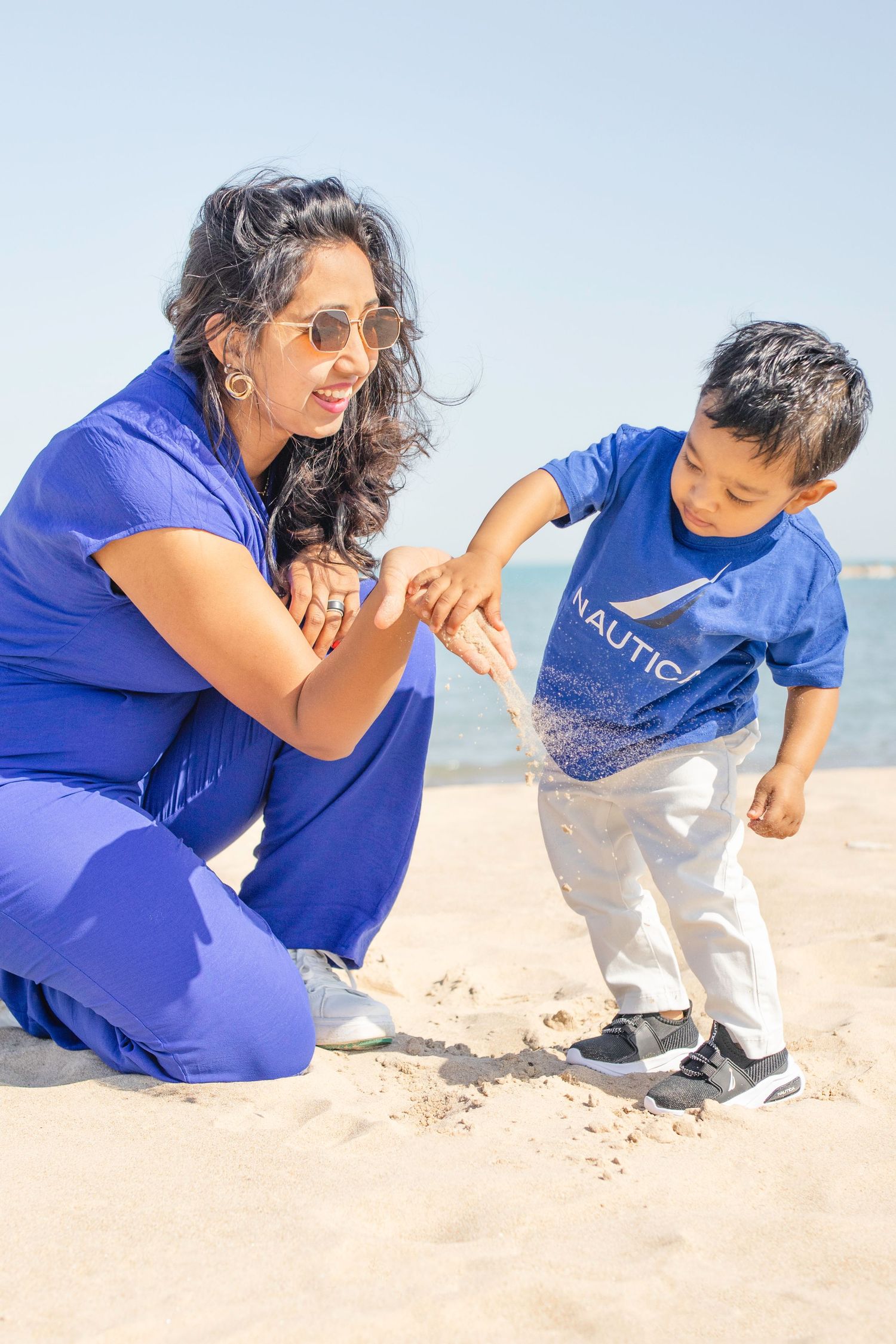 A child and adult explore the beach together, both wearing matching blue outfits as they look at something on the sand.