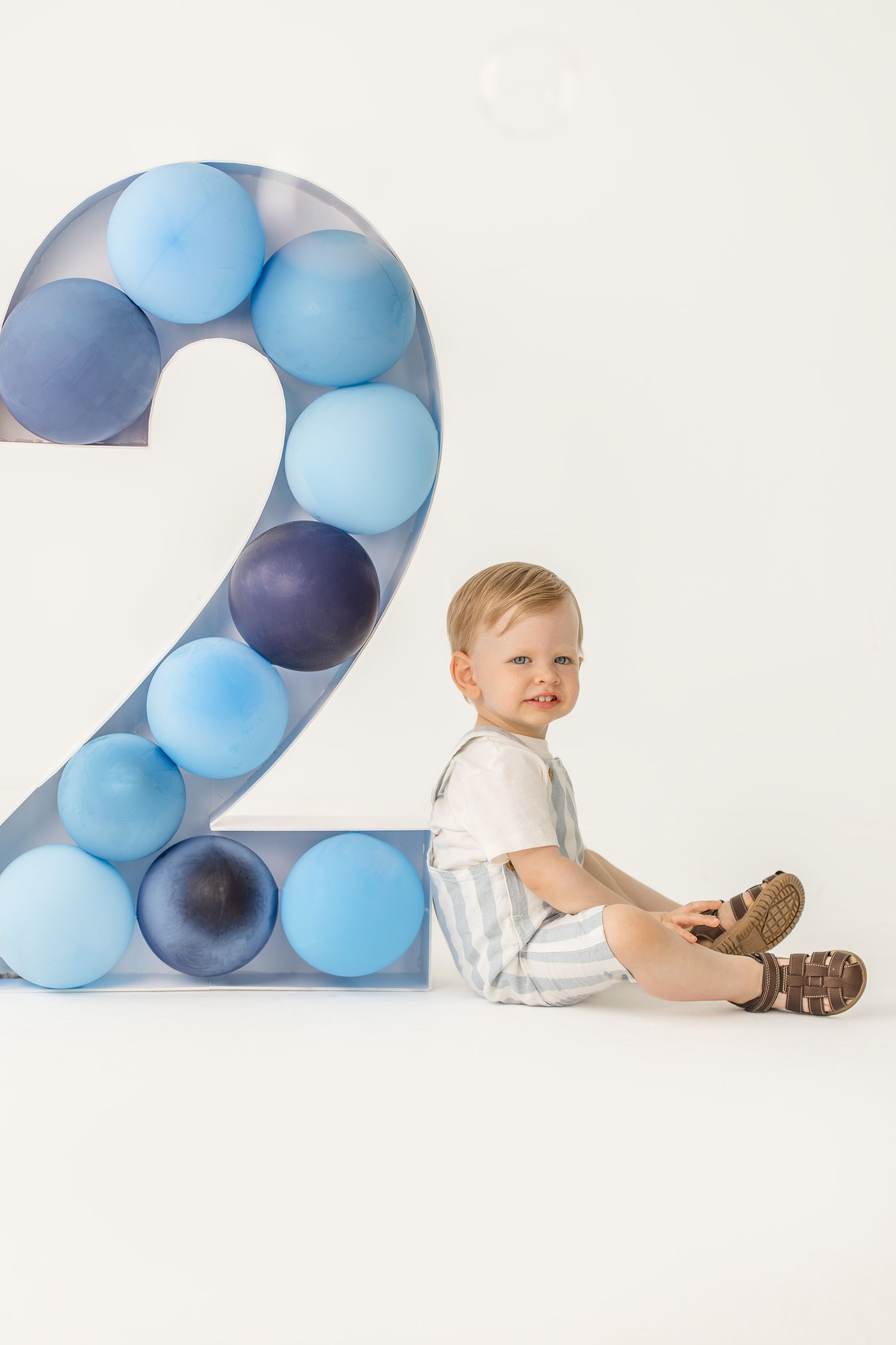 Toddler boy in striped overalls sitting on the floor next to a large number 2 filled with blue balloons.