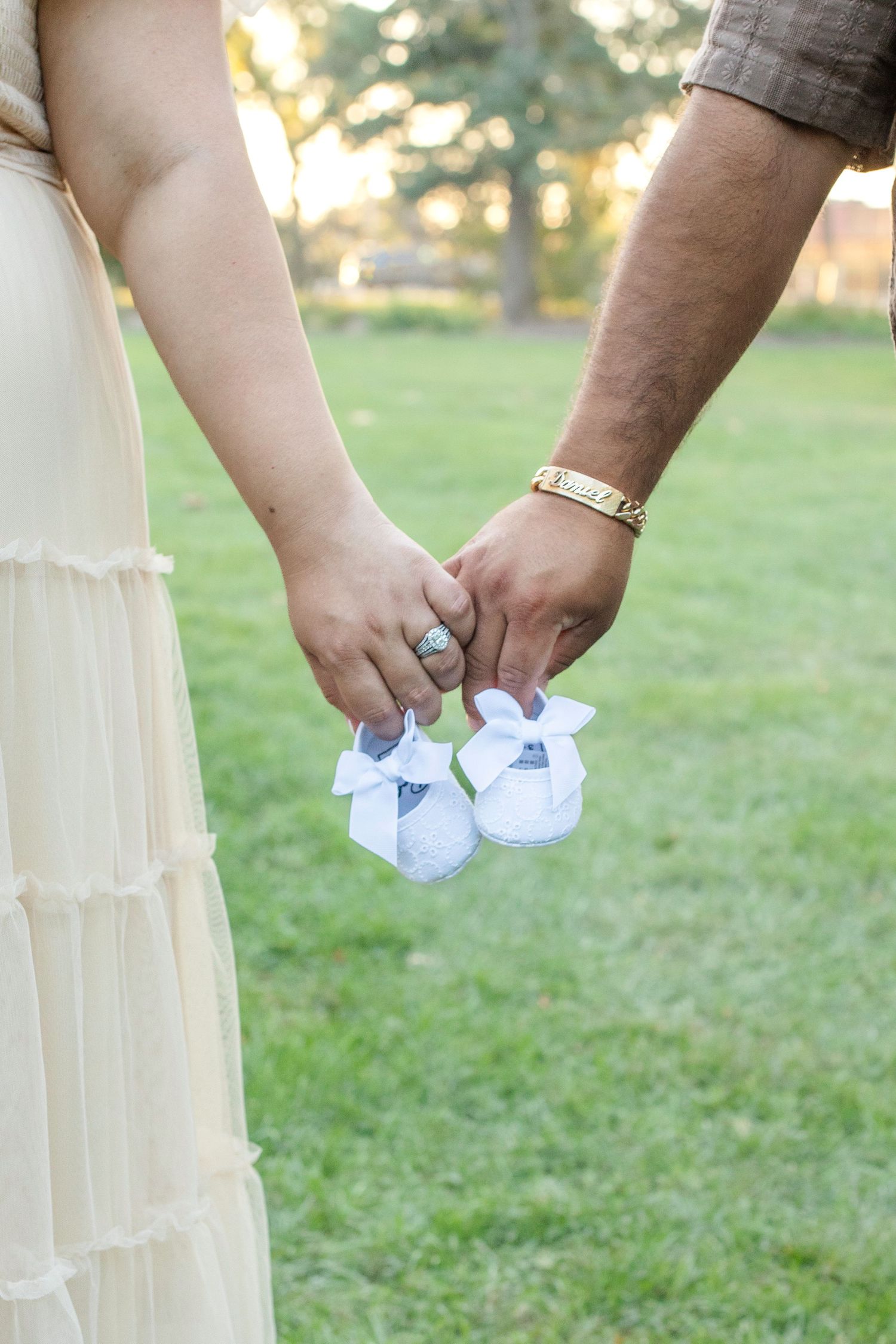 A pair of hands holds tiny white baby booties against a soft green grassy background in golden sunlight.