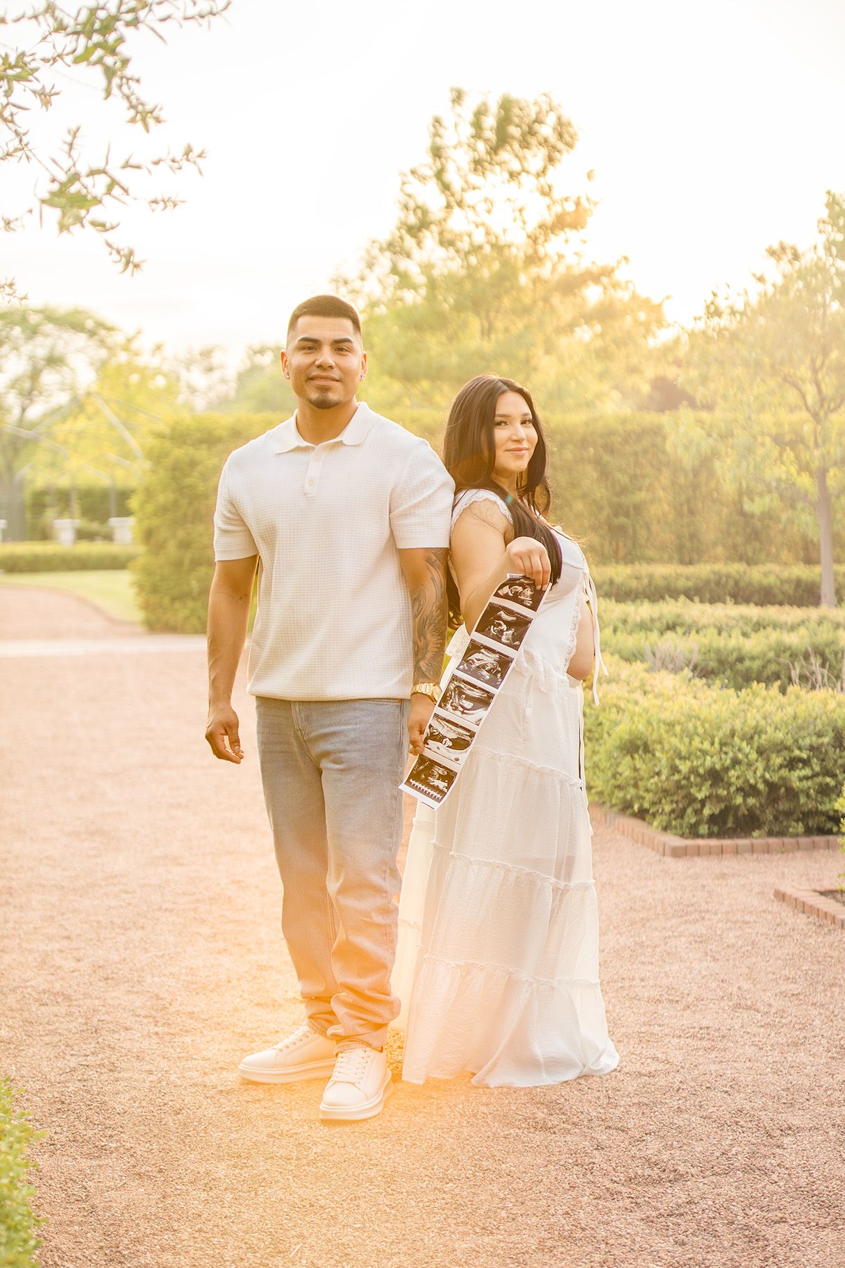 Pregnant woman in white dress and partner walk a garden path holding ultrasound strips during golden hour maternity shoot.