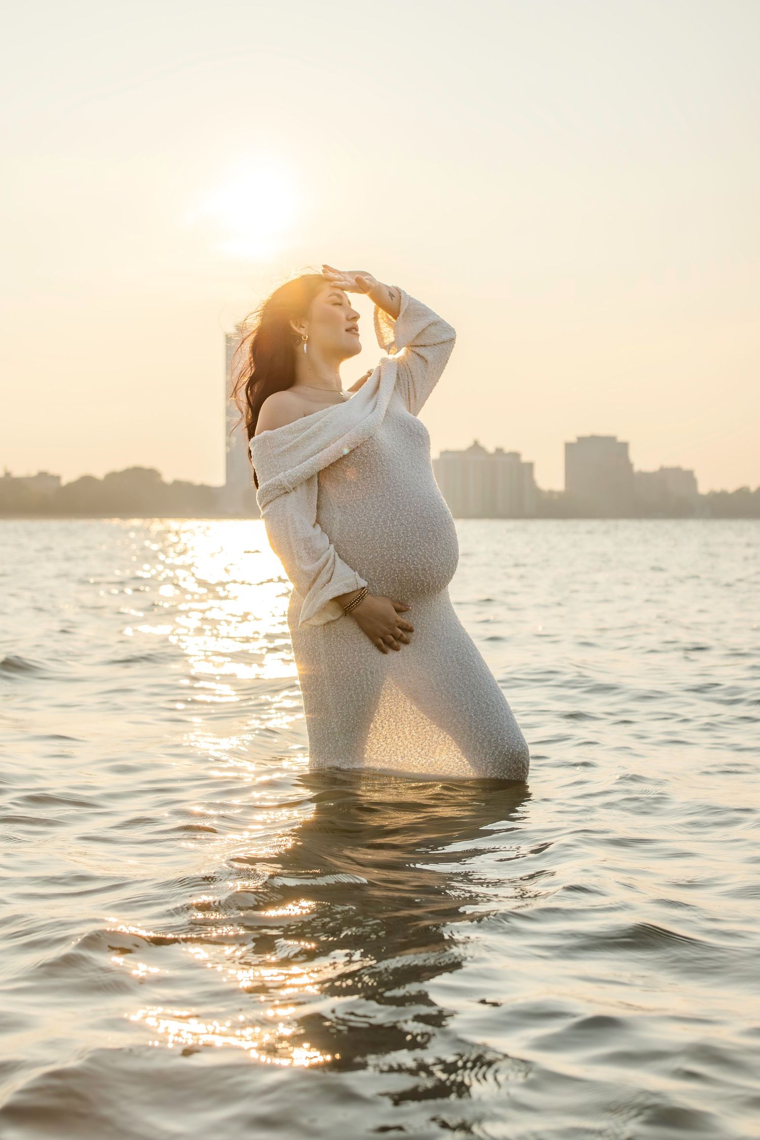 A pregnant woman in a white dress stands in calm water during golden hour with a city skyline in the background.
