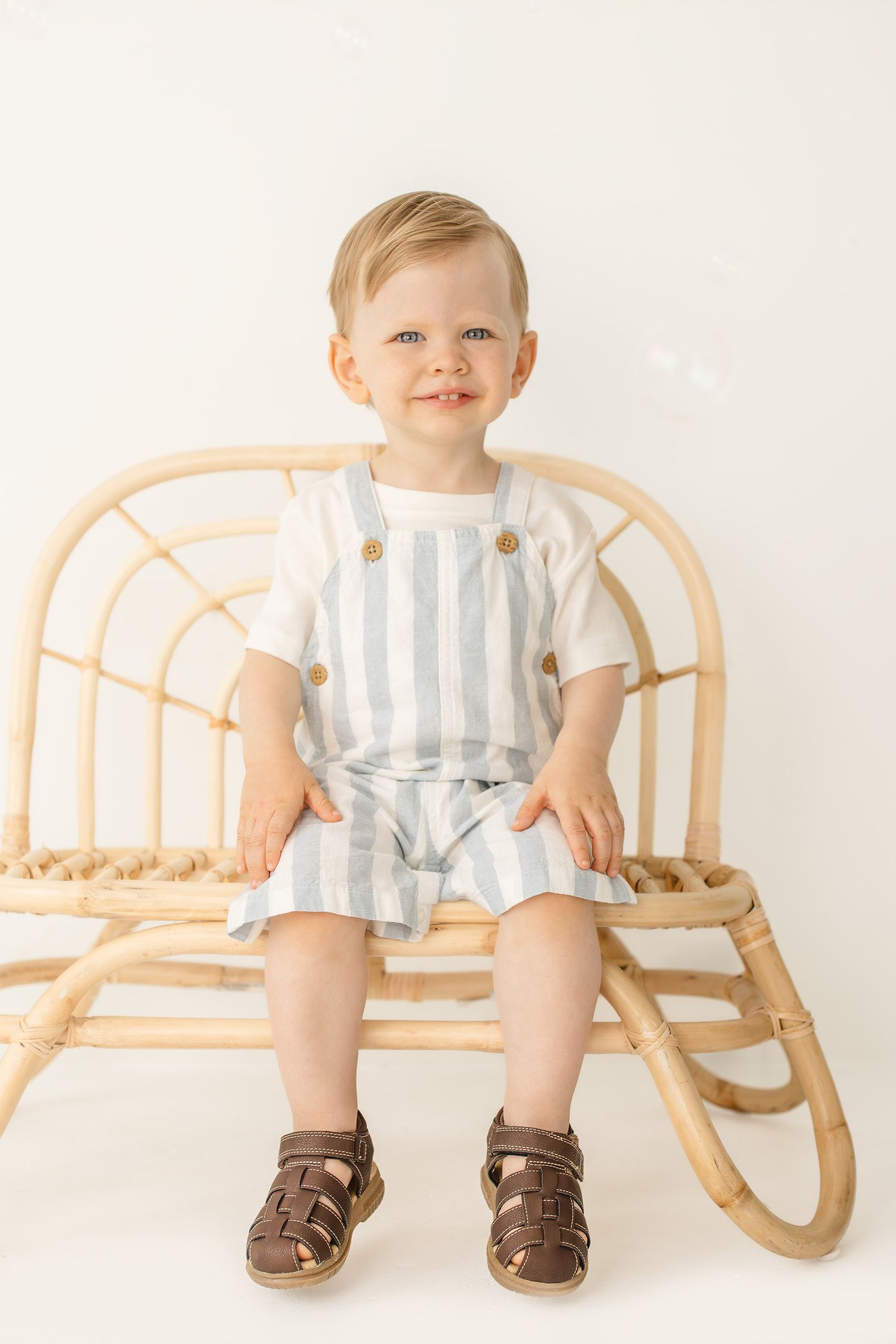 Smiling toddler boy in striped shortall overalls sitting on a rattan bench against a white background.