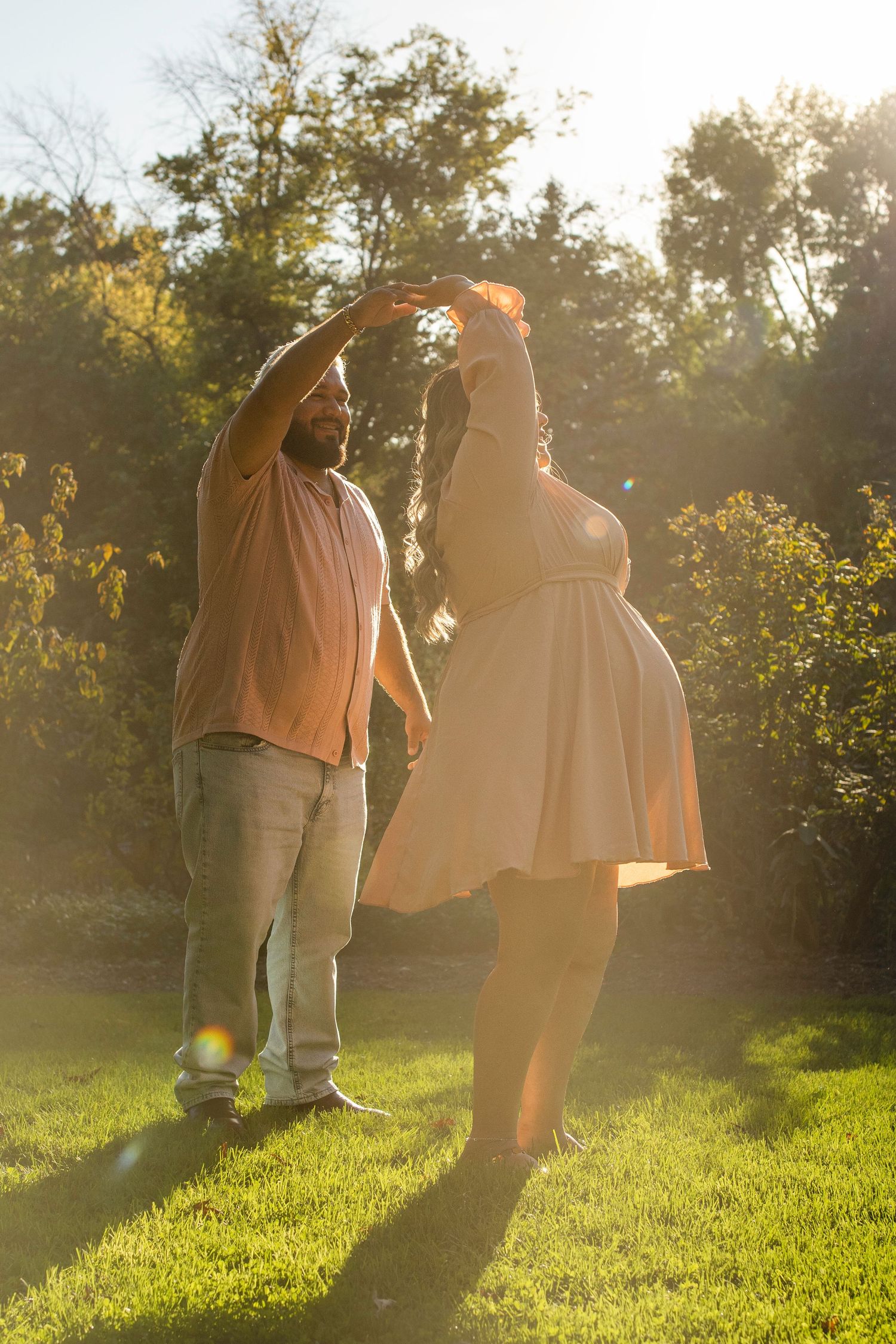 A couple dances together on a grassy hill during golden hour with sunlight streaming through trees.