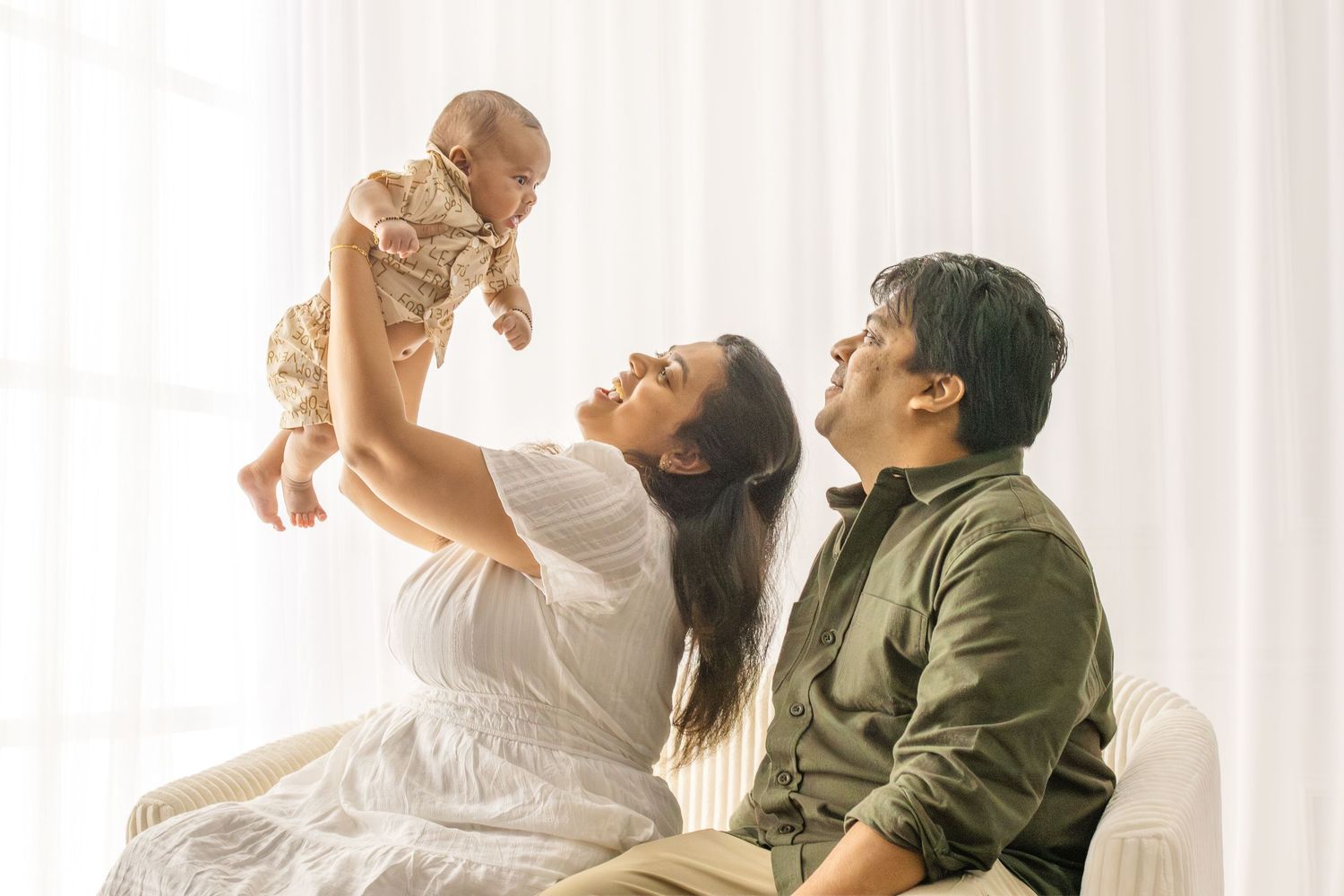 Parents in olive green and white outfits playfully lift and interact with their baby against a bright window background.