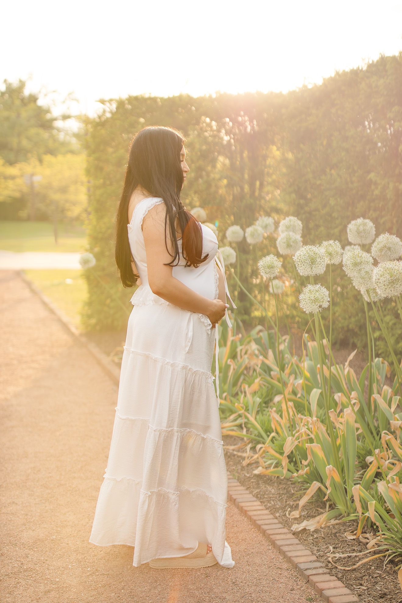 Woman in white tiered dress standing in sunlit garden with allium flowers blooming at golden hour.