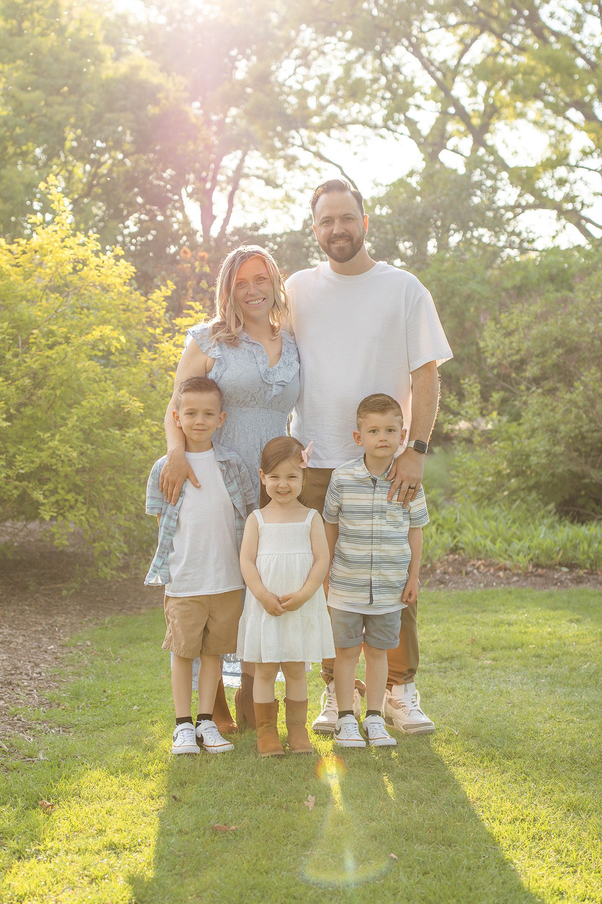 Family of five posing outdoors in golden sunlight, two adults and three young children smiling on green grass surrounded by trees.