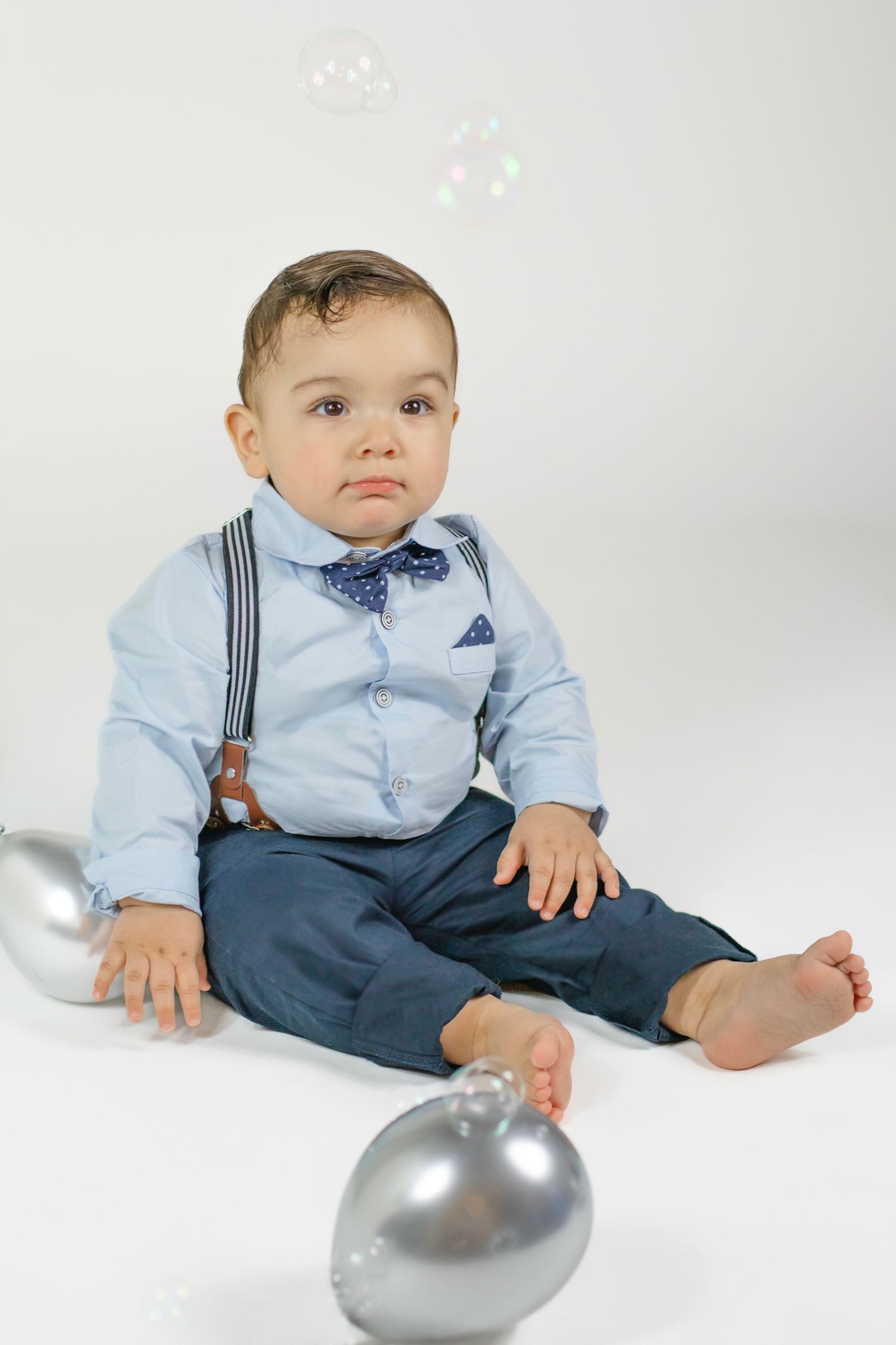 Adorable baby boy in blue shirt, bow tie, suspenders, and navy pants sits barefoot with silver ornaments and bubbles.