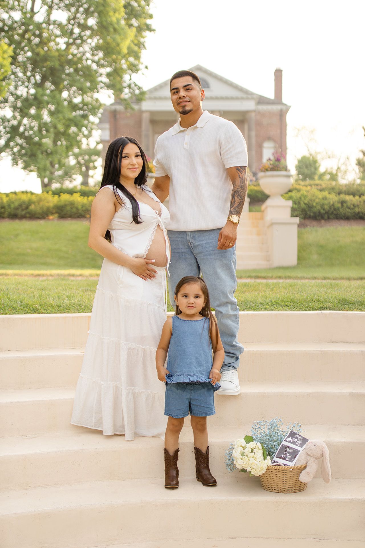Pregnant woman in white dress poses with partner and toddler daughter on steps during golden hour maternity photo session.