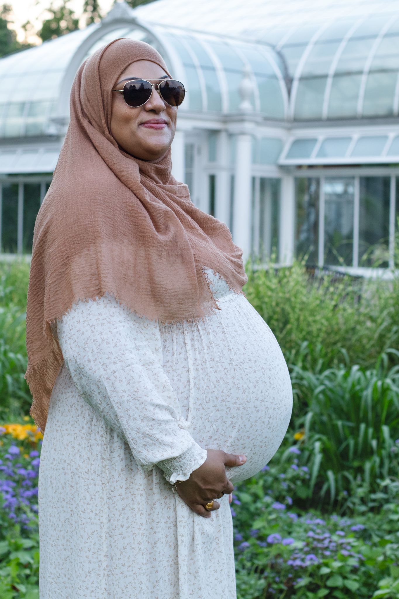 Pregnant woman in hijab and white dress smiles while standing near greenhouse.