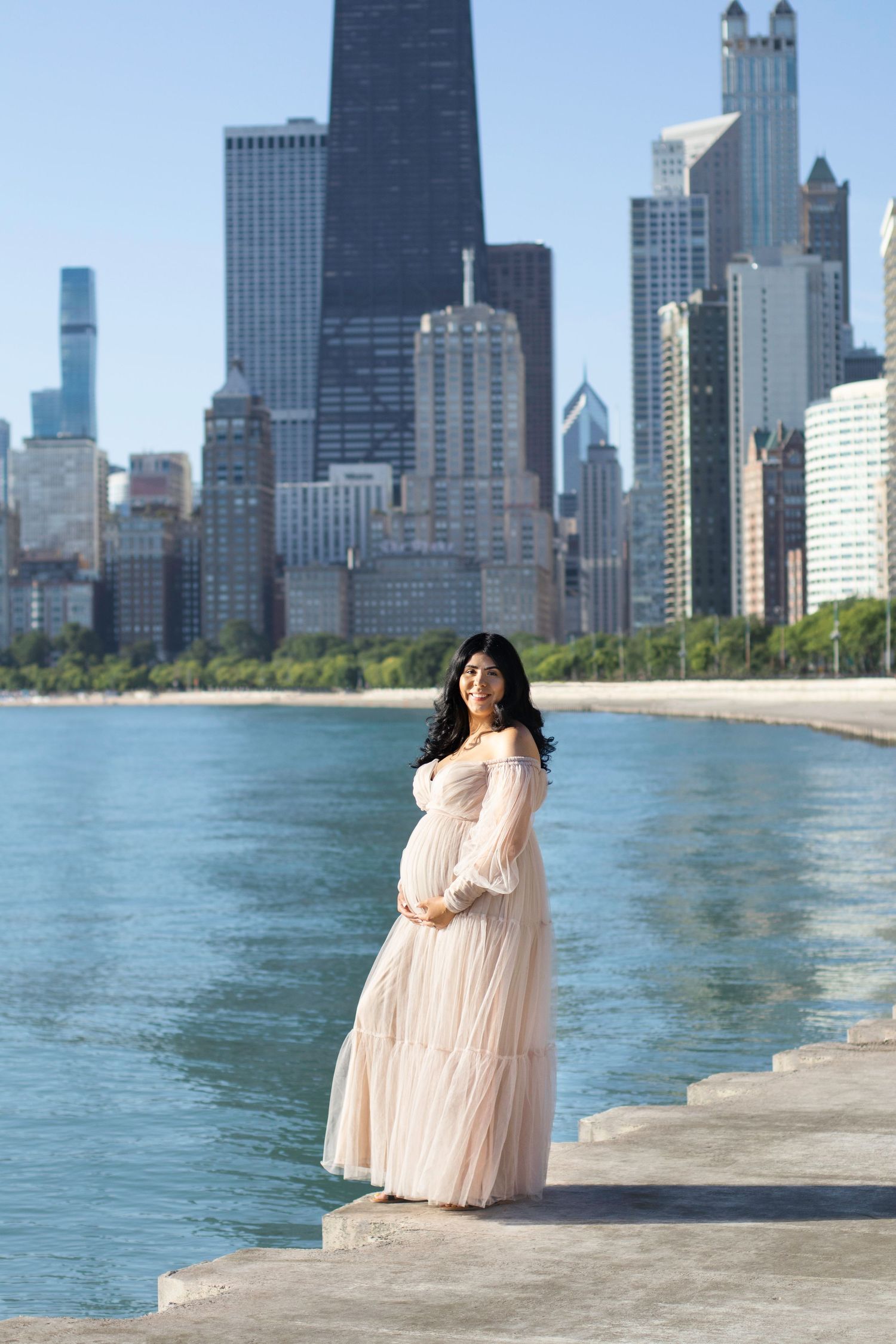 Maternity photo in flowing beige dress against Chicago skyline and Lake Michigan.