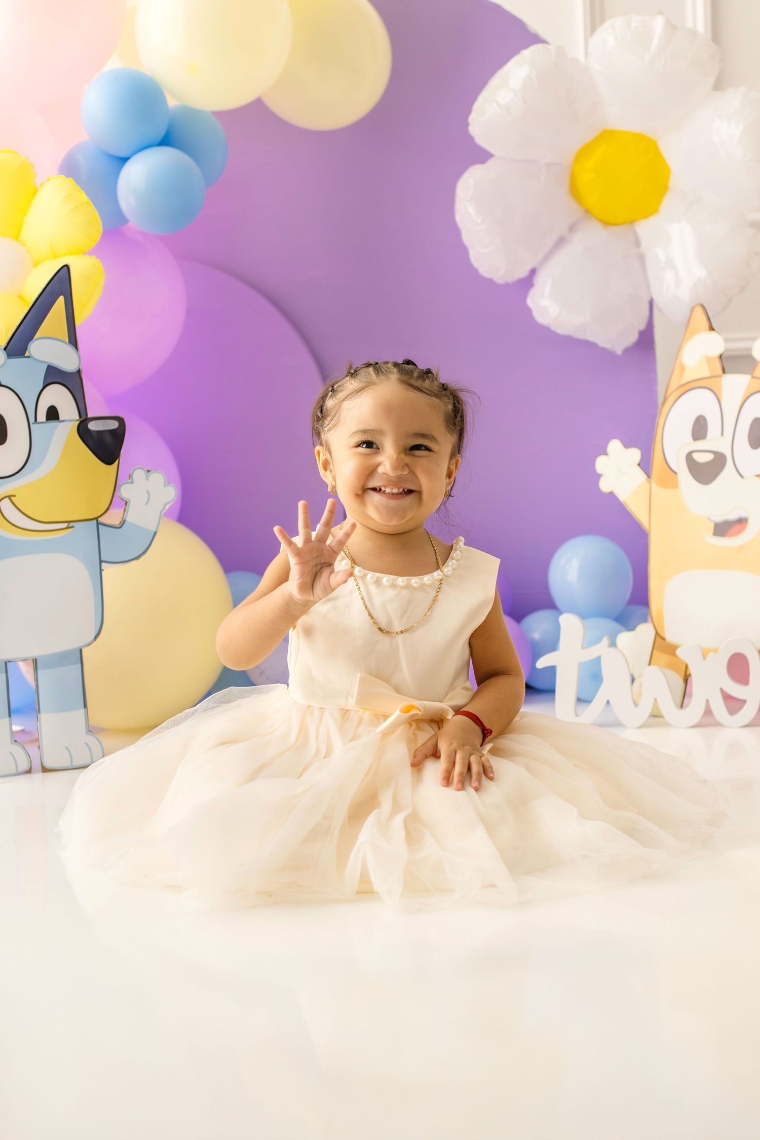 Happy toddler girl in white tulle dress waves at a Bluey-themed 2nd birthday party with colorful balloon decorations.