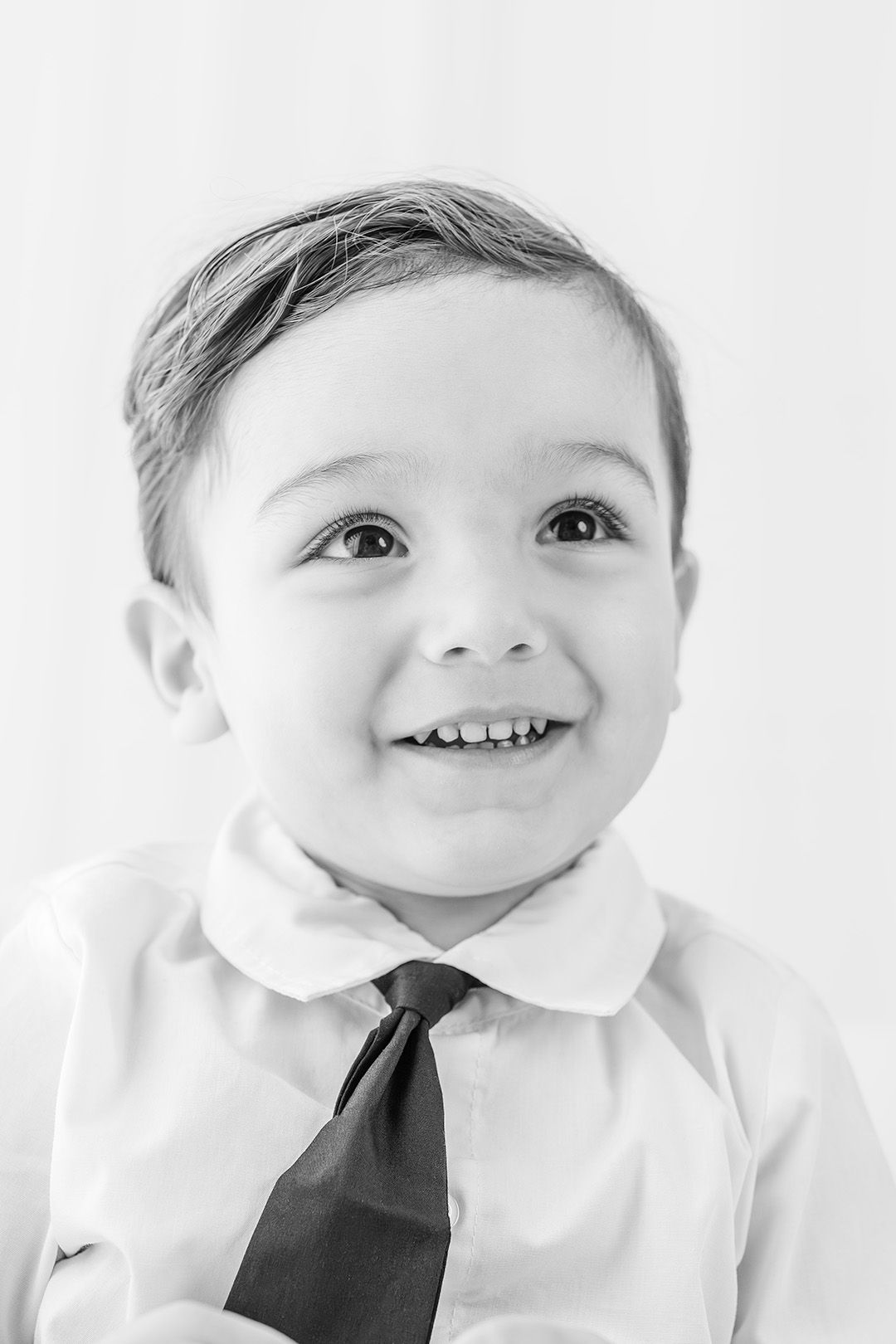 Black and white portrait of a smiling young boy wearing a white dress shirt and tie, looking up cheerfully.