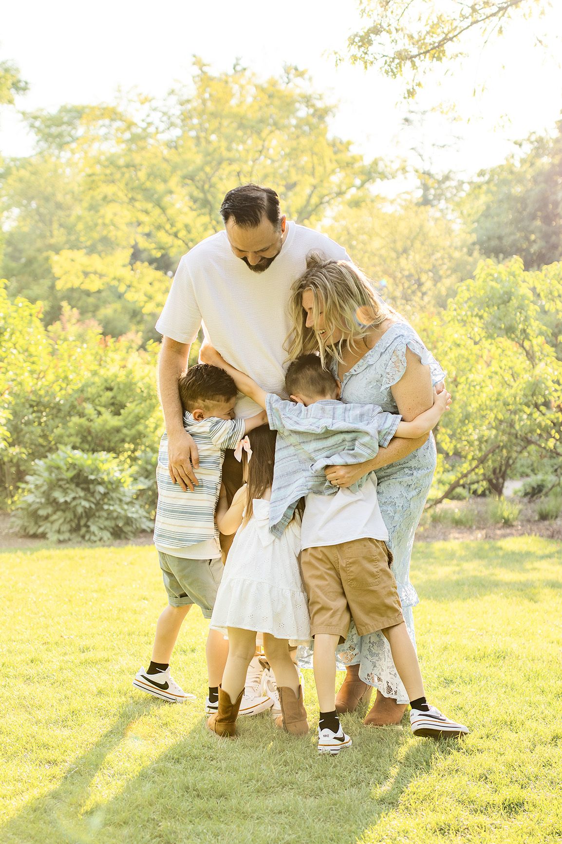 Family of five sharing a warm group hug outdoors on green grass, surrounded by sunlit trees.