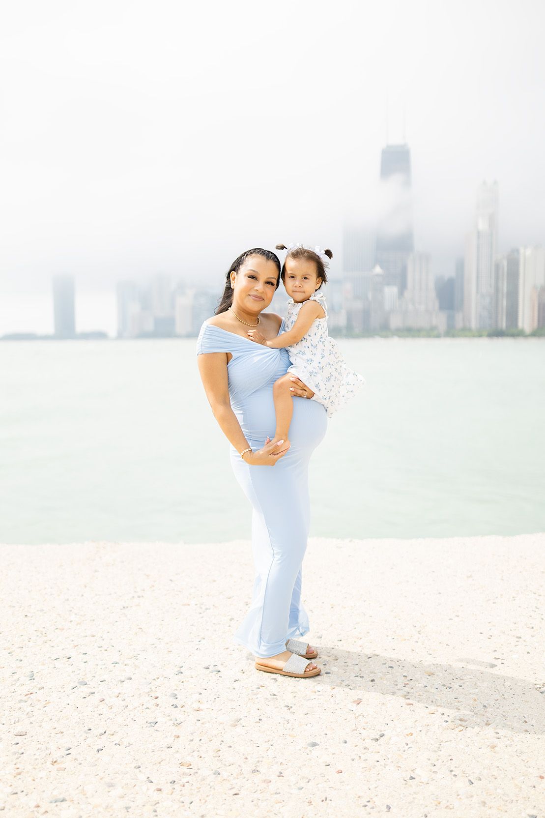Pregnant woman in light blue off-shoulder dress holds toddler girl near Chicago lakefront skyline.