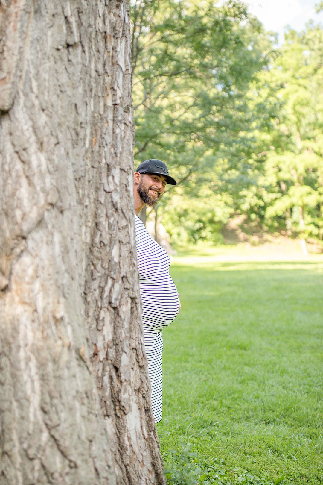 Smiling bearded man in striped shirt and cap peeking out from behind a large tree trunk in a sunny park.