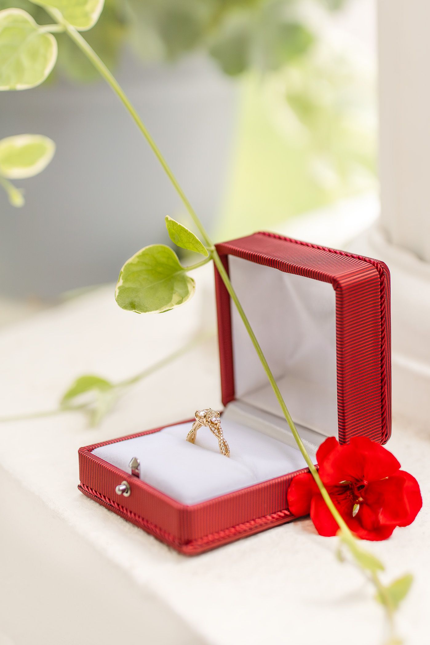 Gold diamond engagement ring in open red jewelry box with red flower and green leaves on white surface.