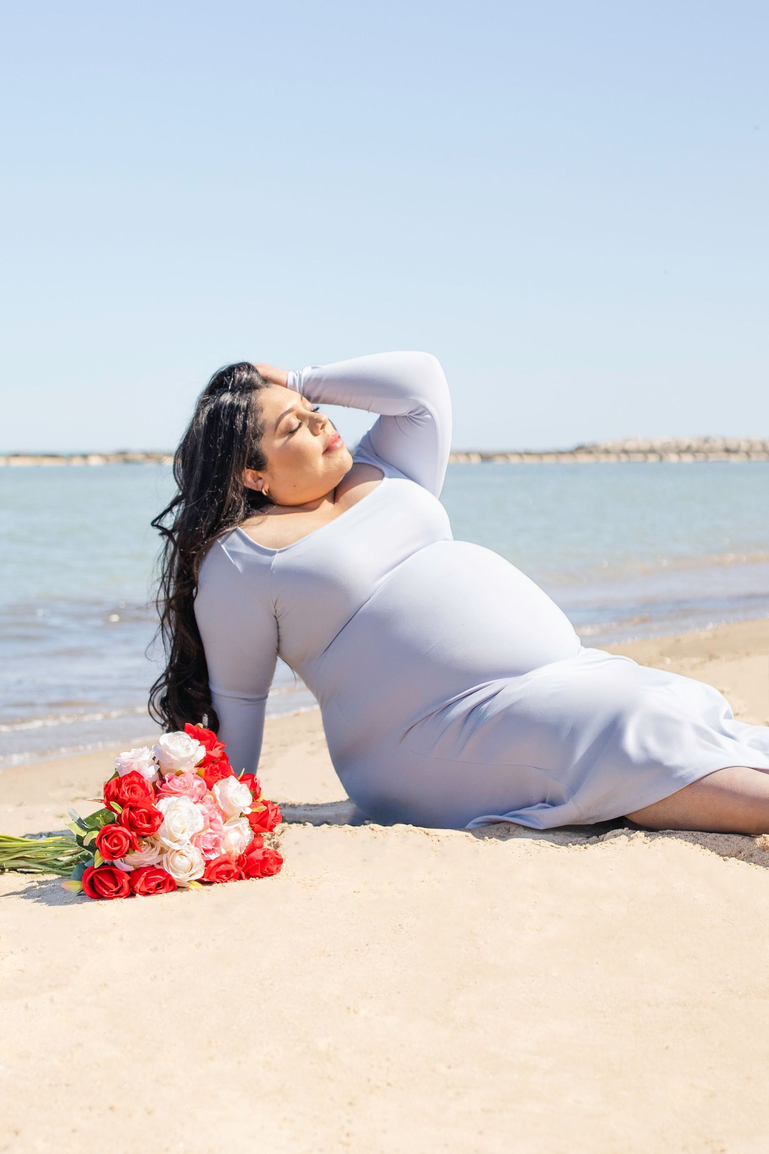A person in a white dress reclines on a beach with red and pink roses beside them as they enjoy the ocean view.